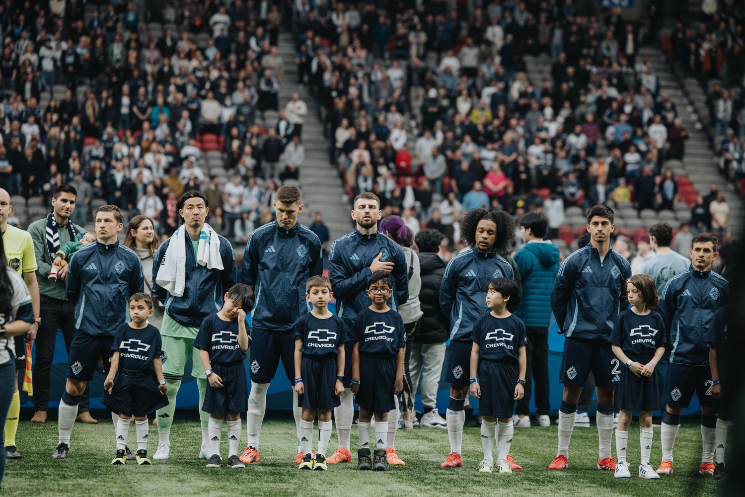 Sports team and youth players lined up on field before large stadium crowd.