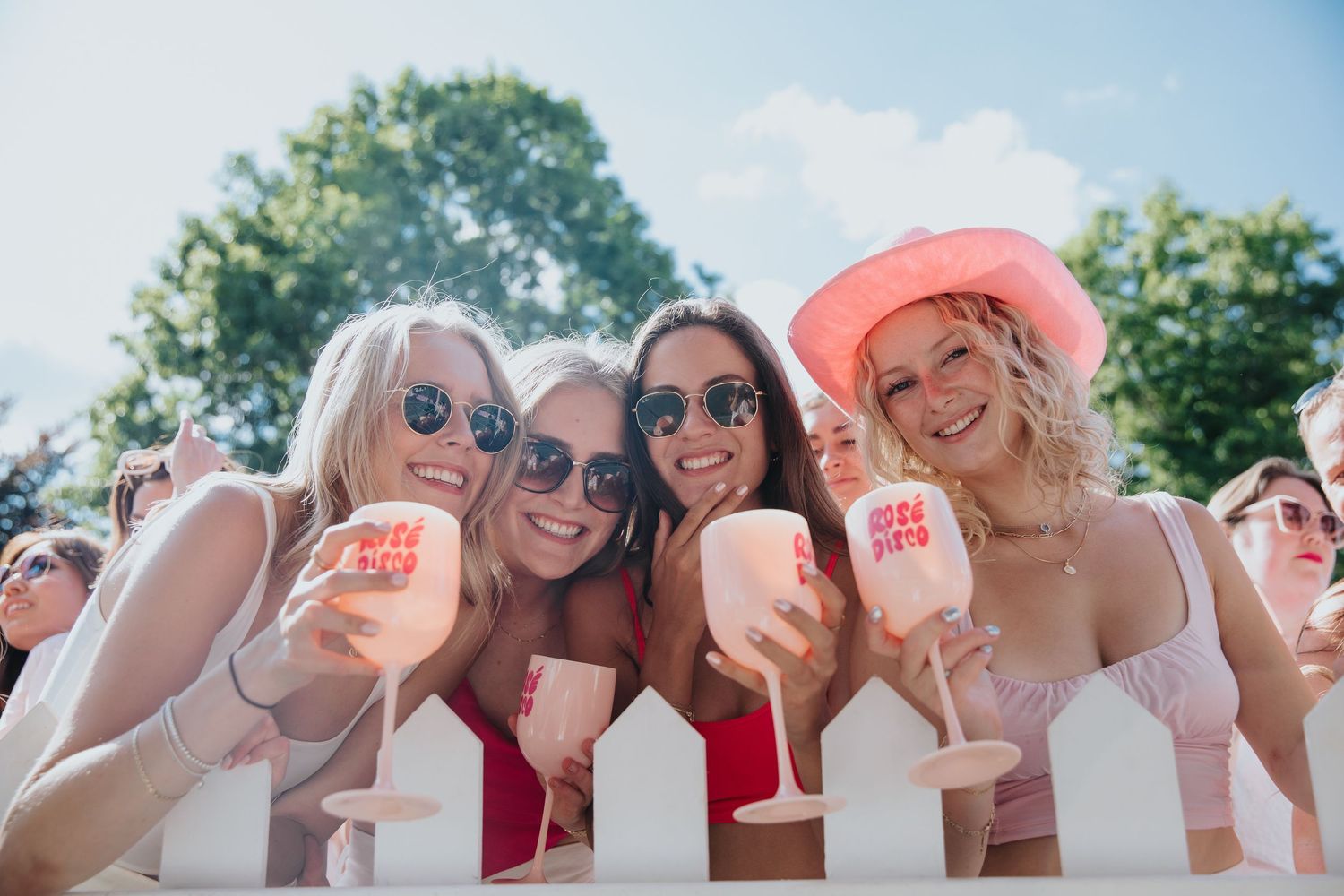 Four smiling women in pink outfits holding branded cups at a sunny outdoor festival event.