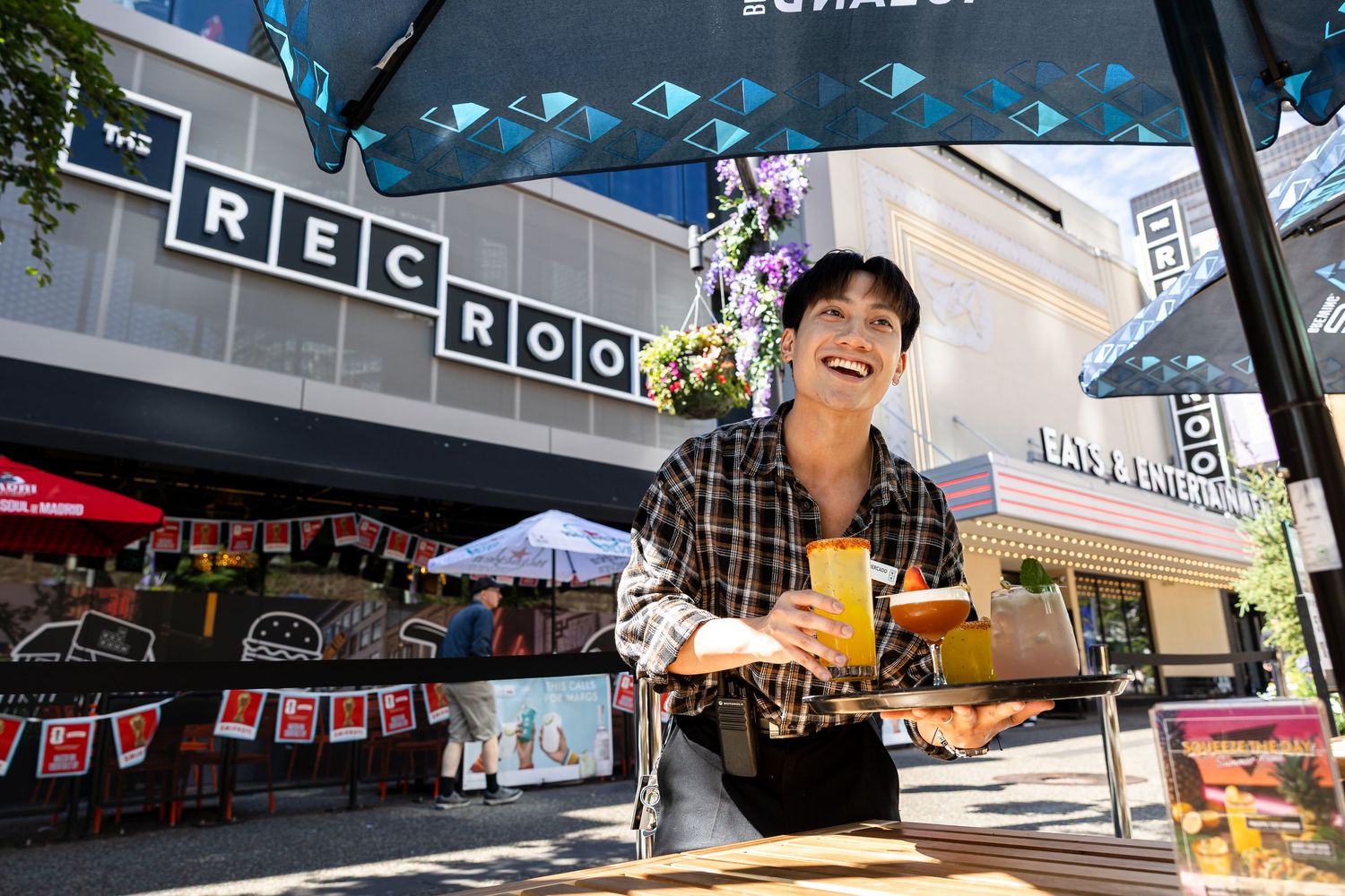 A smiling person holds drinks and food outside The Rec Room restaurant on a sunny day.