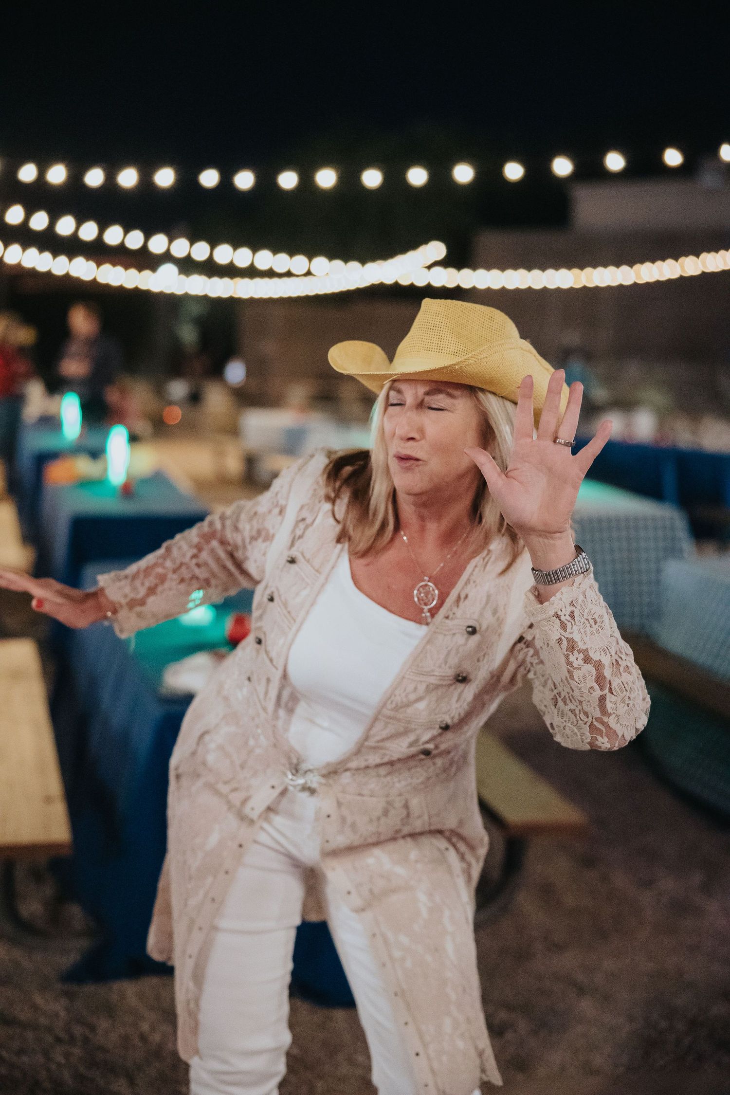 Older woman in a yellow cowboy hat and white lace outfit dancing outdoors at a night event with string lights.