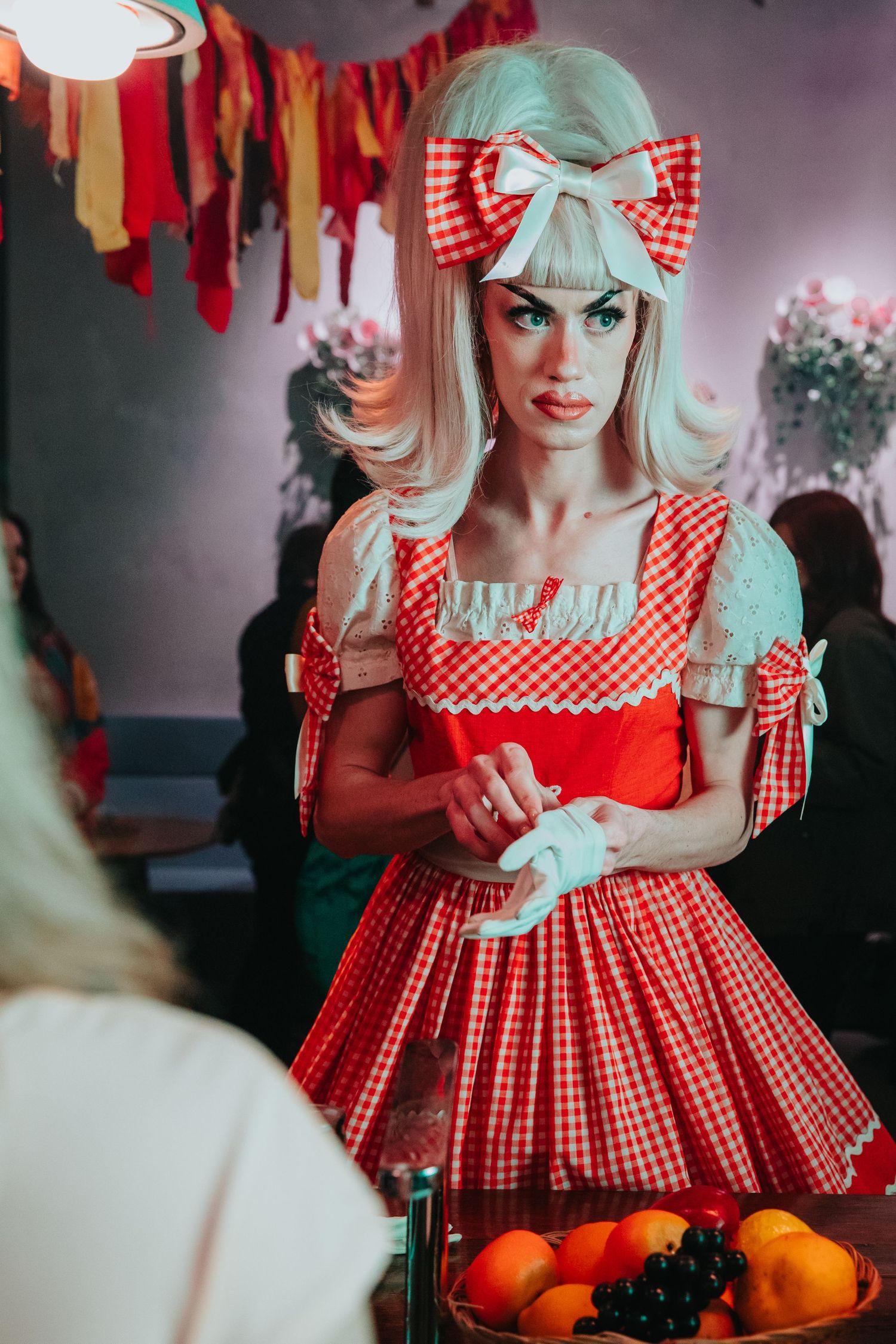 Woman in red gingham dress and blonde wig with bow poses near fruit display at themed party.