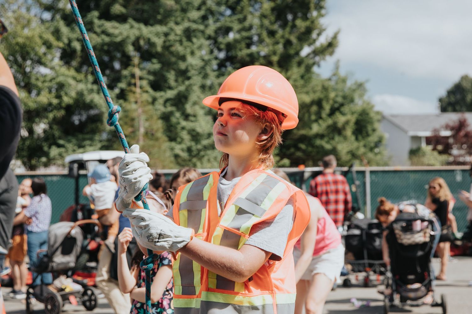 Woman in orange hard hat and safety vest holds a rope at an outdoor construction-themed event on a sunny day.