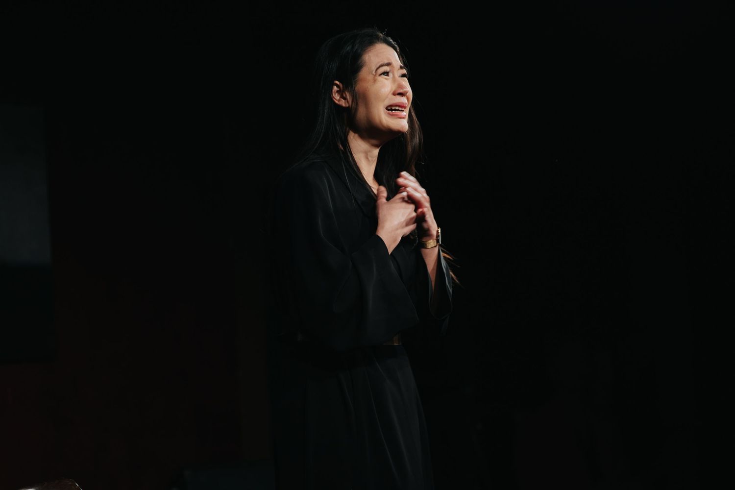 Asian woman in black outfit smiling on dark stage with hands clasped to chest.