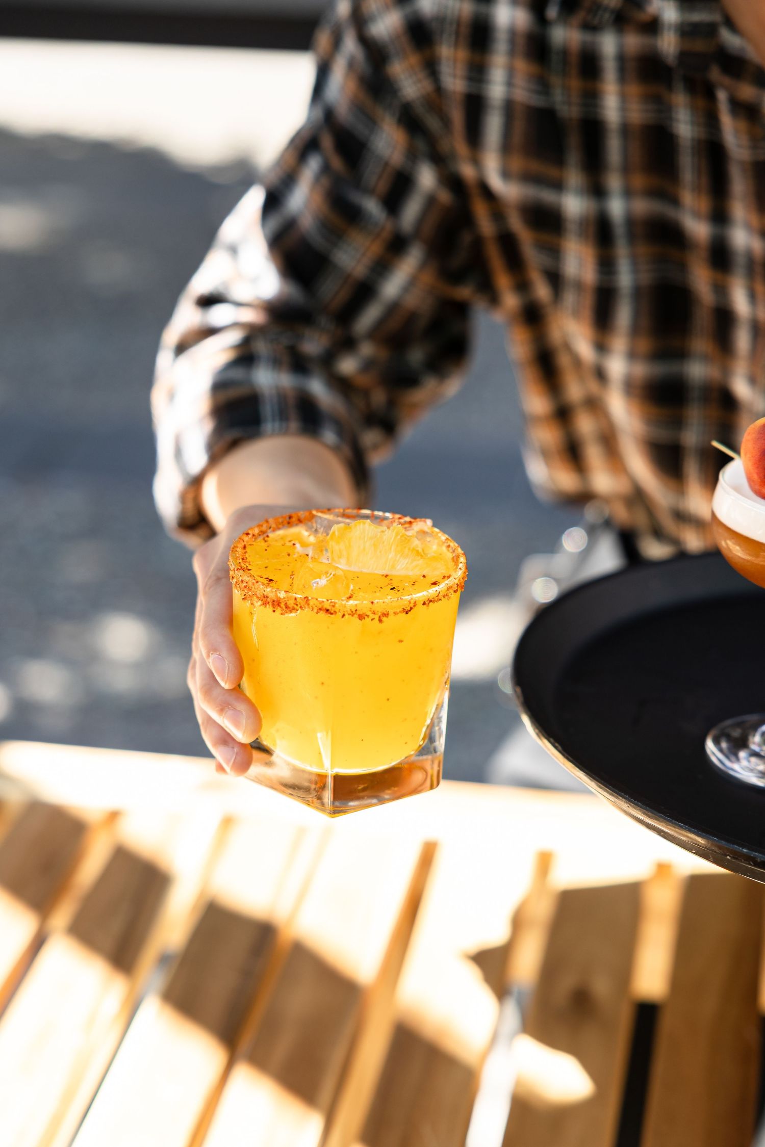 A server places an orange cocktail with a salted rim on a wooden table in bright sunlight.