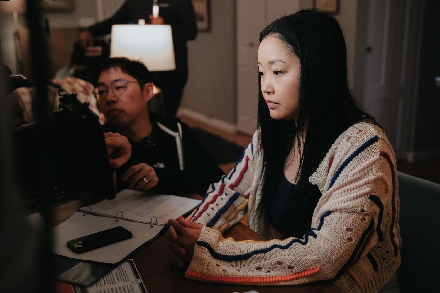 Two students study together at a dimly lit desk, reviewing papers and notes in a cozy home setting.