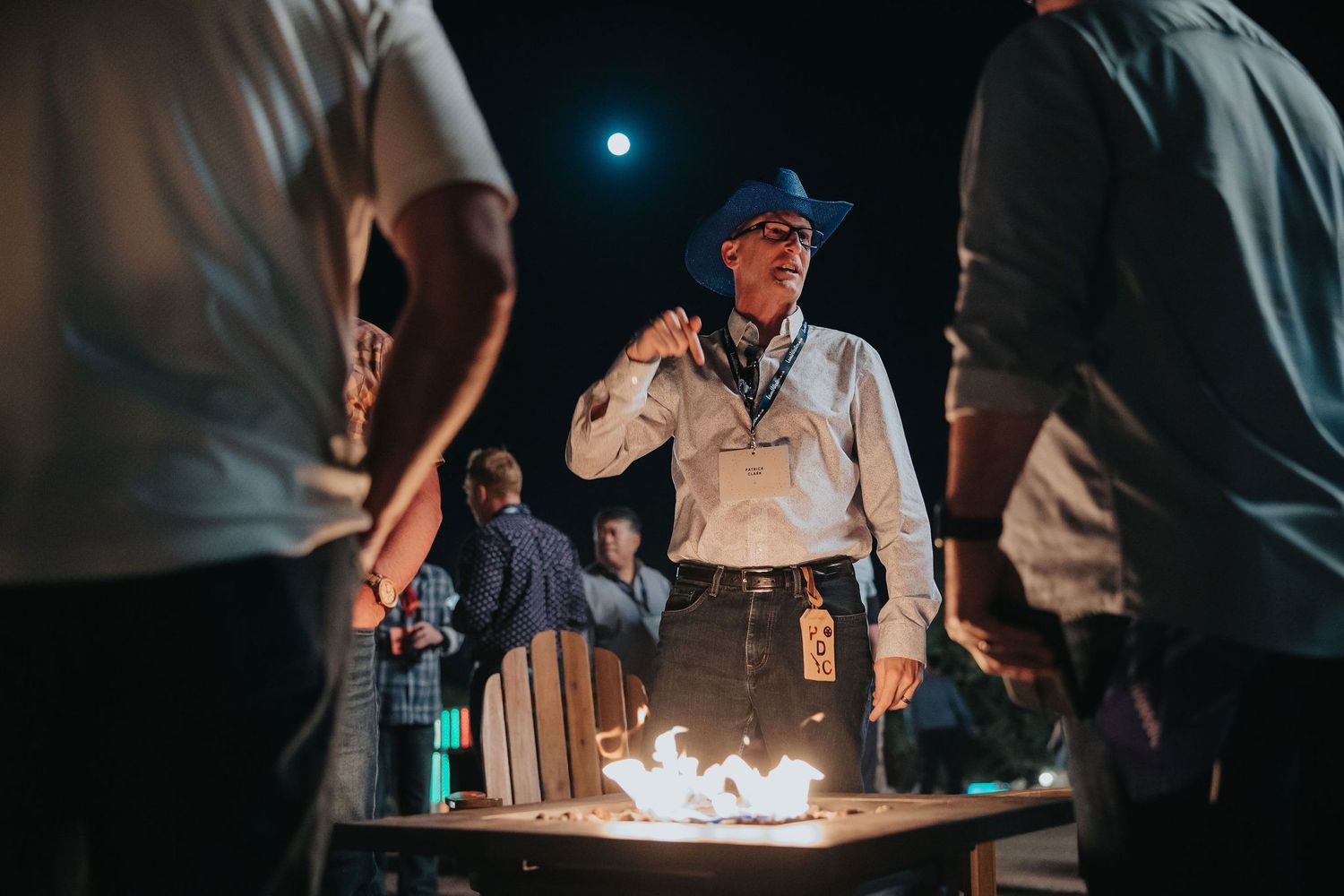 Man in cowboy hat stands by fire pit at night outdoor gathering with guests around him.