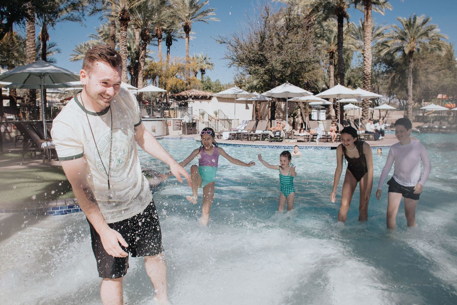 Guests enjoying water activities at an outdoor pool with palm trees and umbrellas.