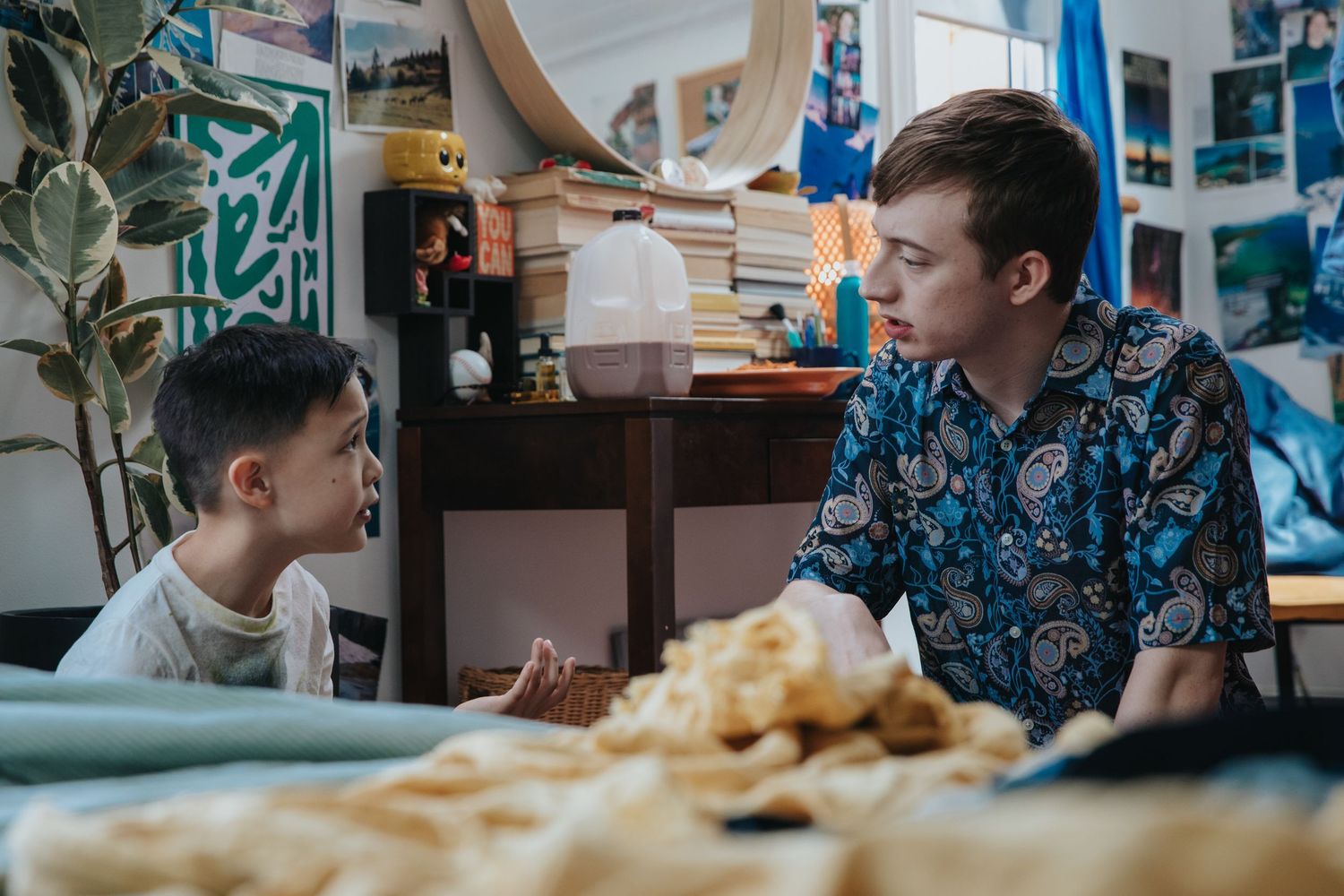 Two people sit at a table covered with food, engaged in conversation in a cozy, art-filled room.