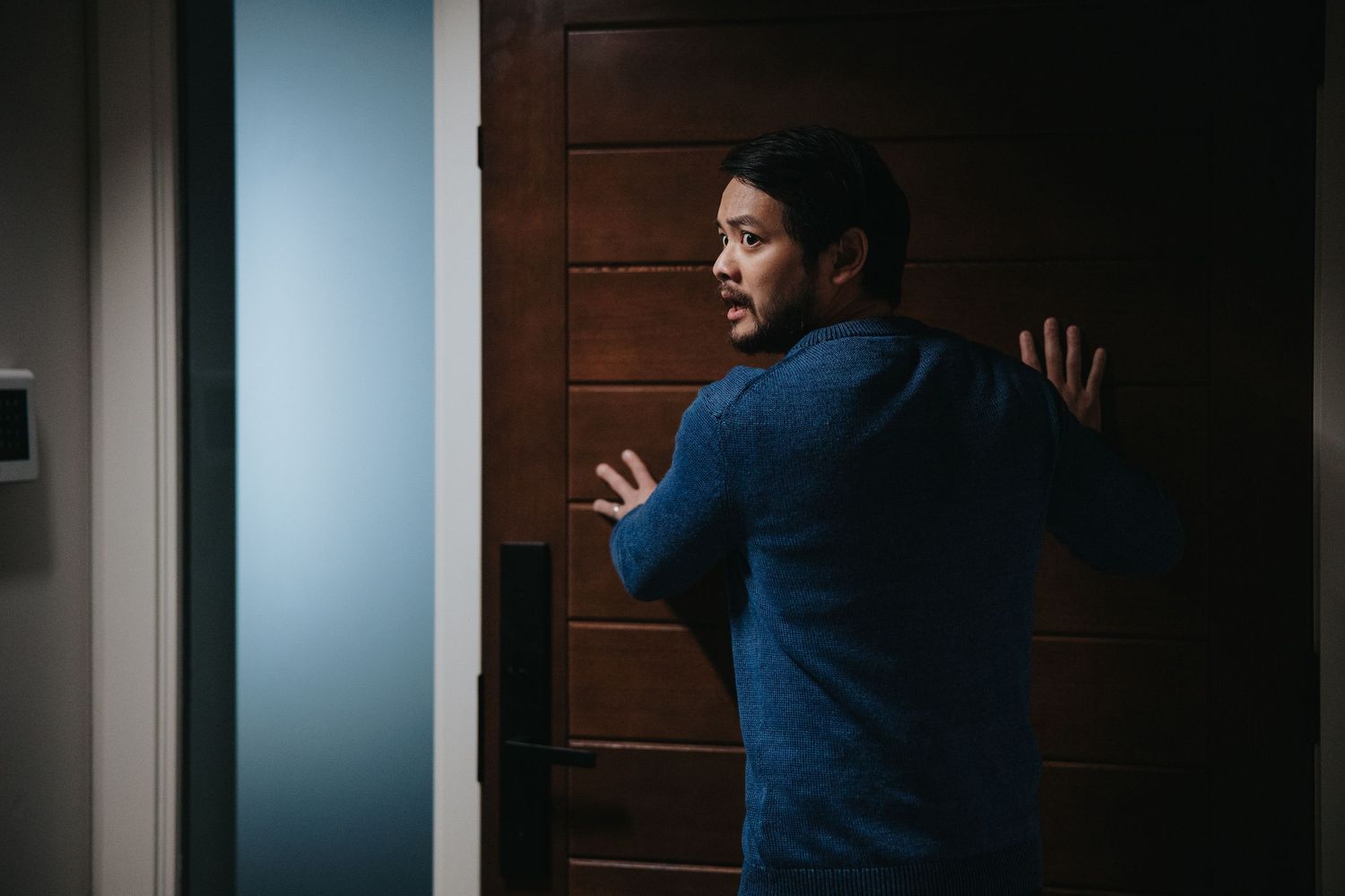 Man in blue sweater cautiously peering around a wooden door in dimly lit hallway.