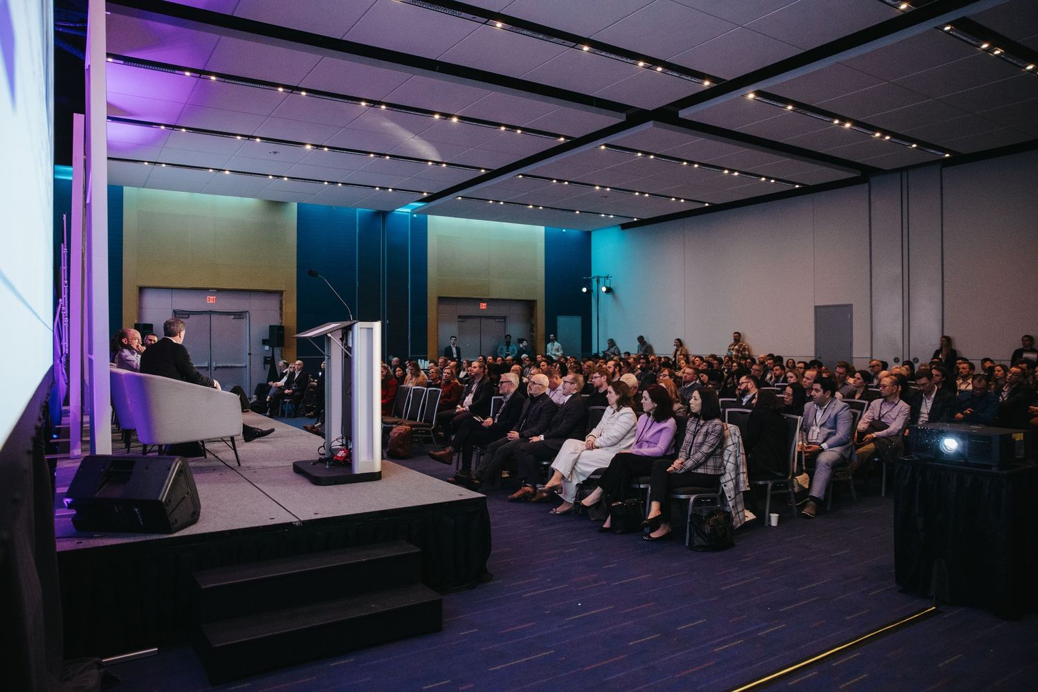 Panoramic view of a conference room with purple lighting and attendees seated in rows facing a stage area.