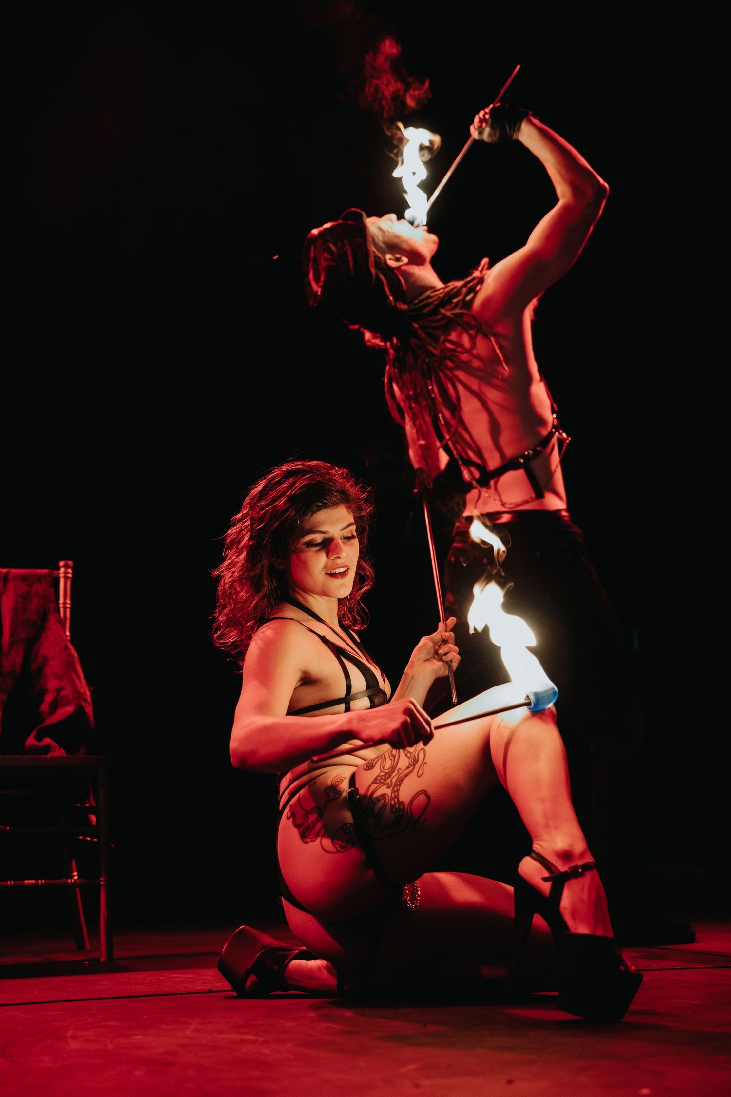 Two fire performers in dramatic red lighting, one seated and one standing, holding flaming batons on stage.