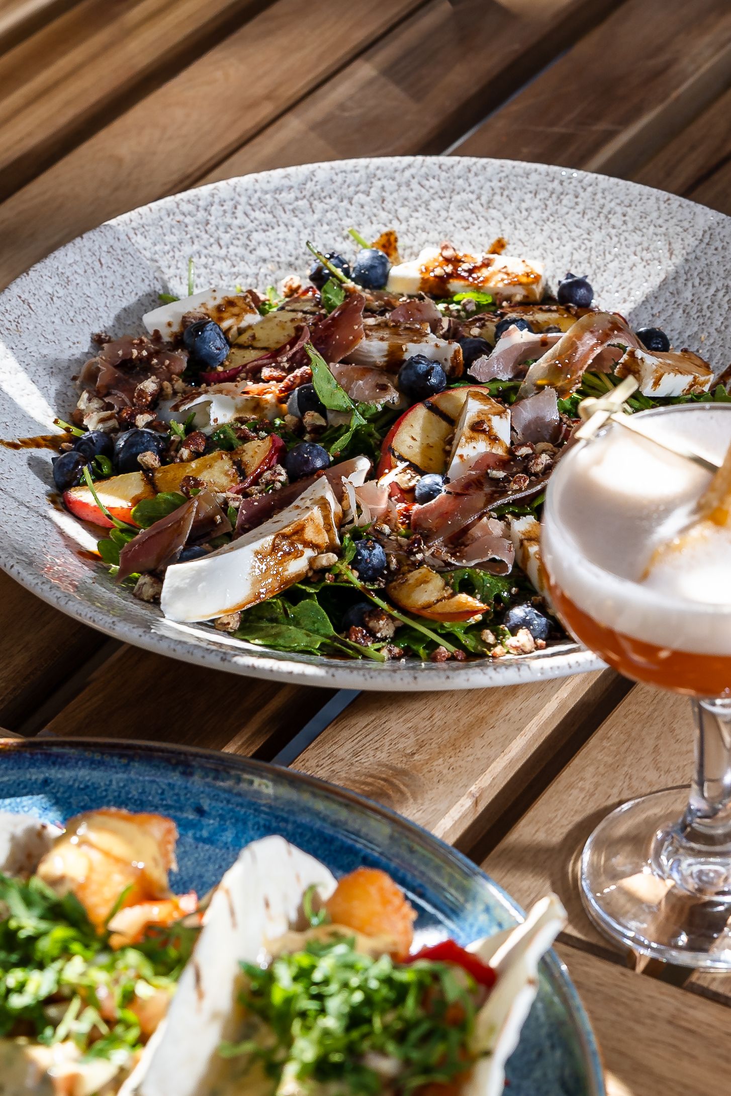 Gourmet salad with prosciutto, blueberries, and greens in a rustic bowl, served alongside a rosé cocktail on a wooden table.