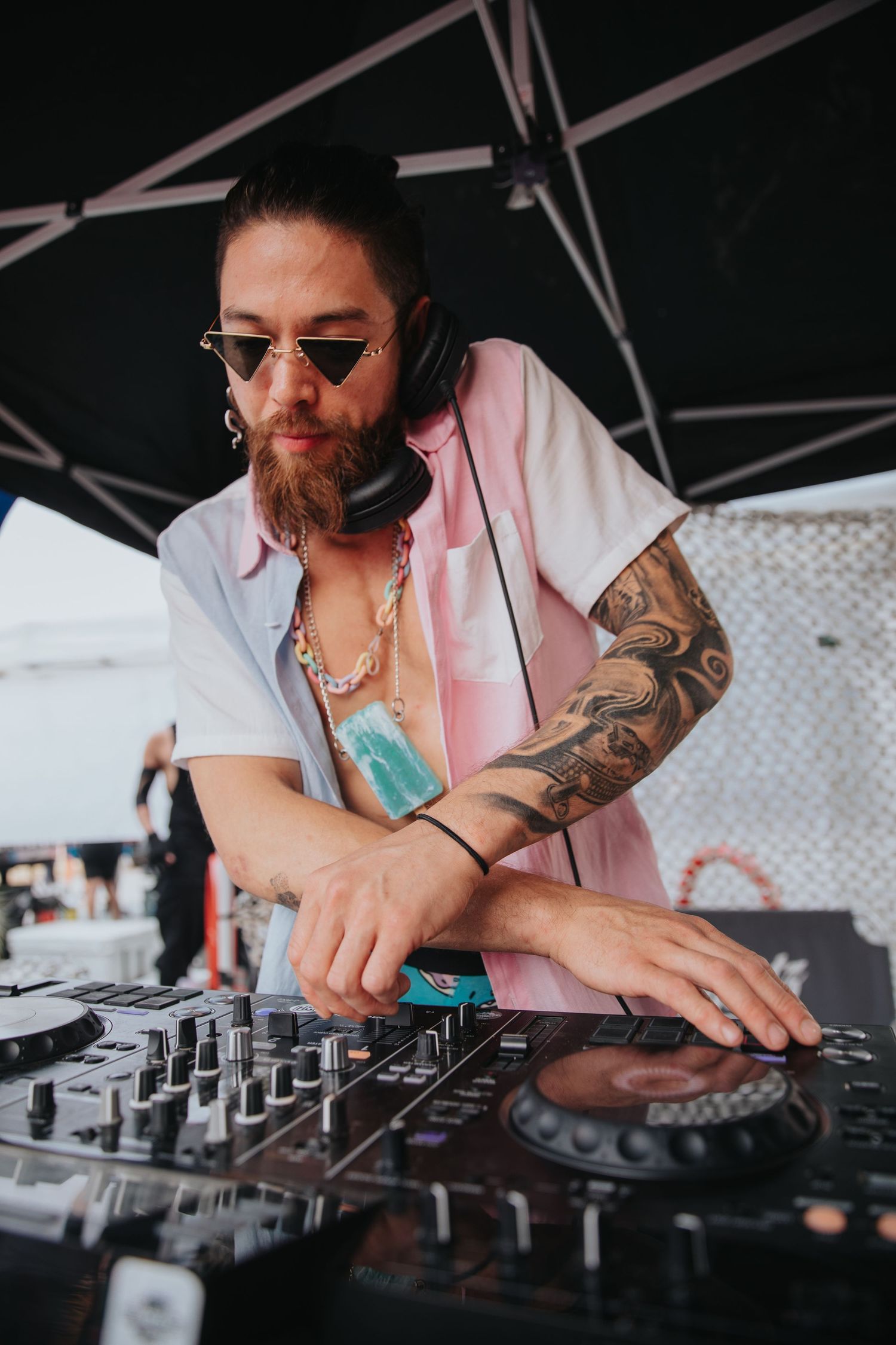Tattooed DJ with sunglasses and headphones mixing music at outdoor festival turntable setup.