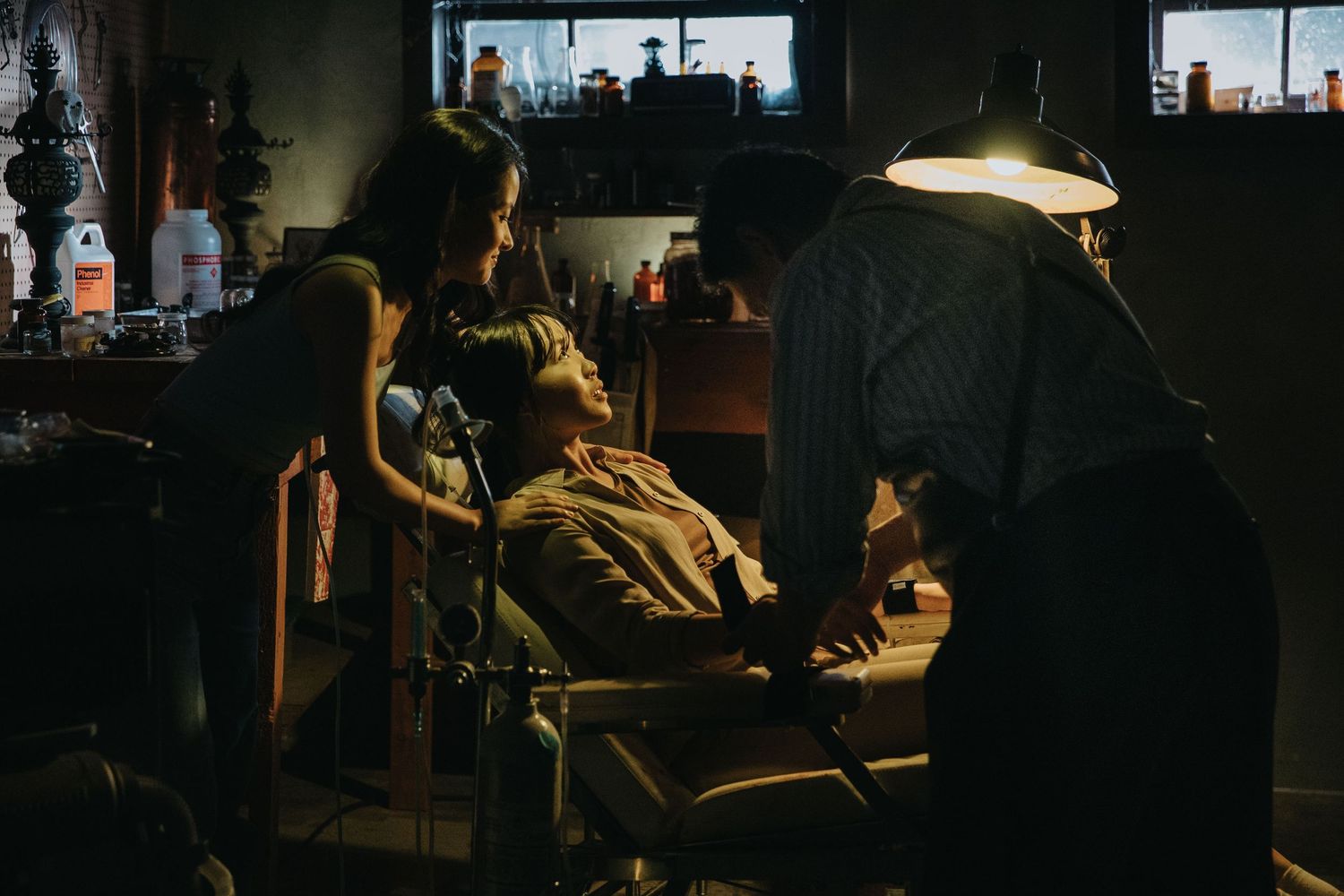 Two people attending to a person reclined in chair under lamp in dark atmospheric room.