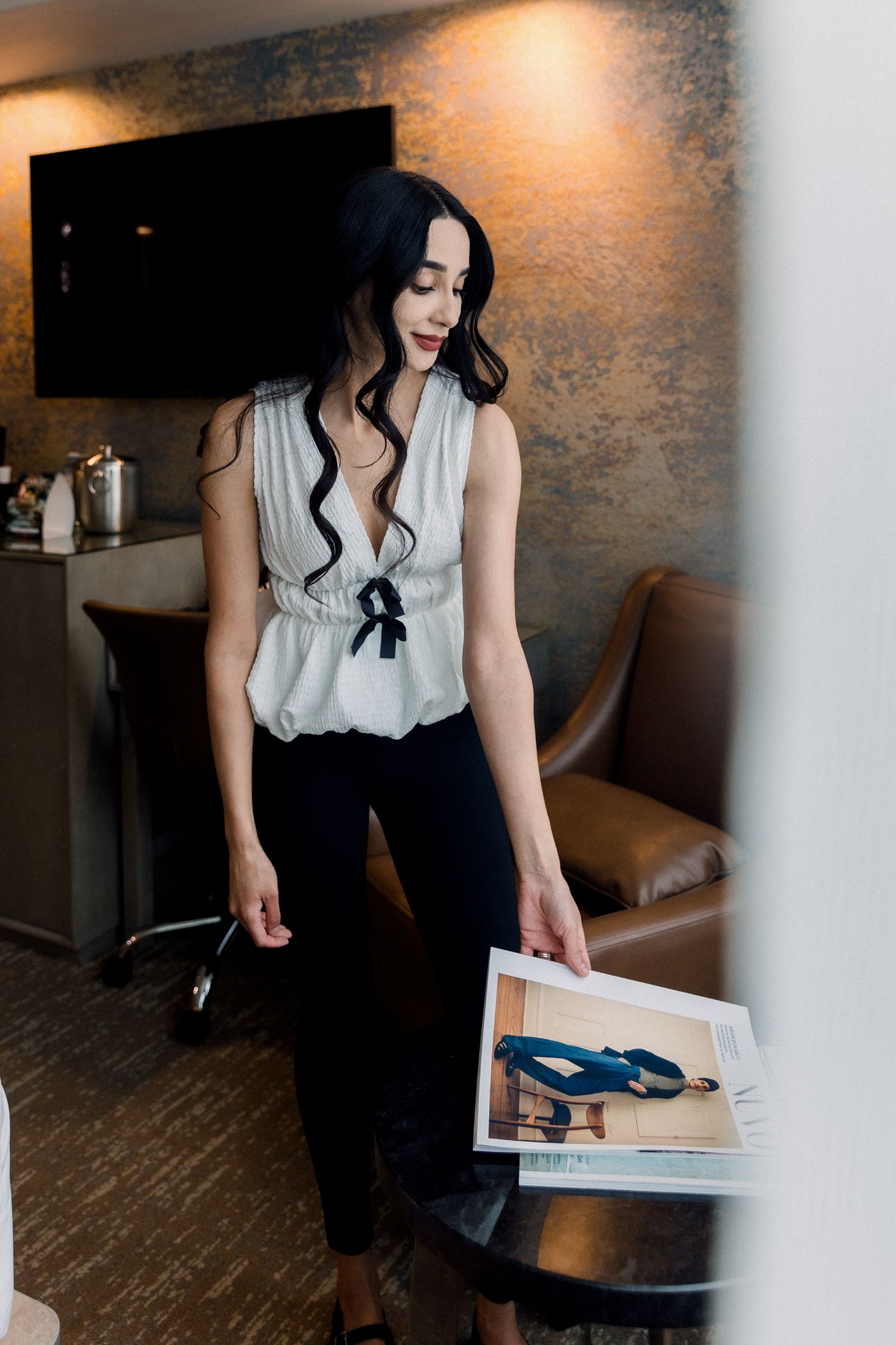 Woman in white peplum top and black pants holds a magazine in a modern hotel room.