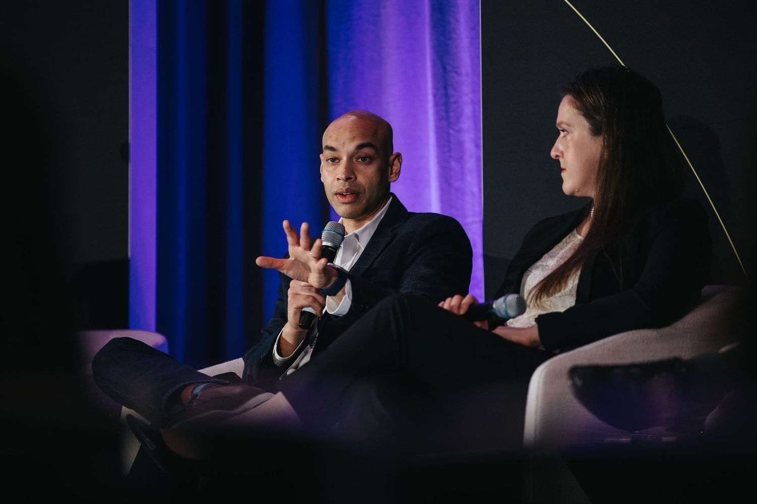 People in conversation on stage at a business conference with purple lighting and dark backdrop.