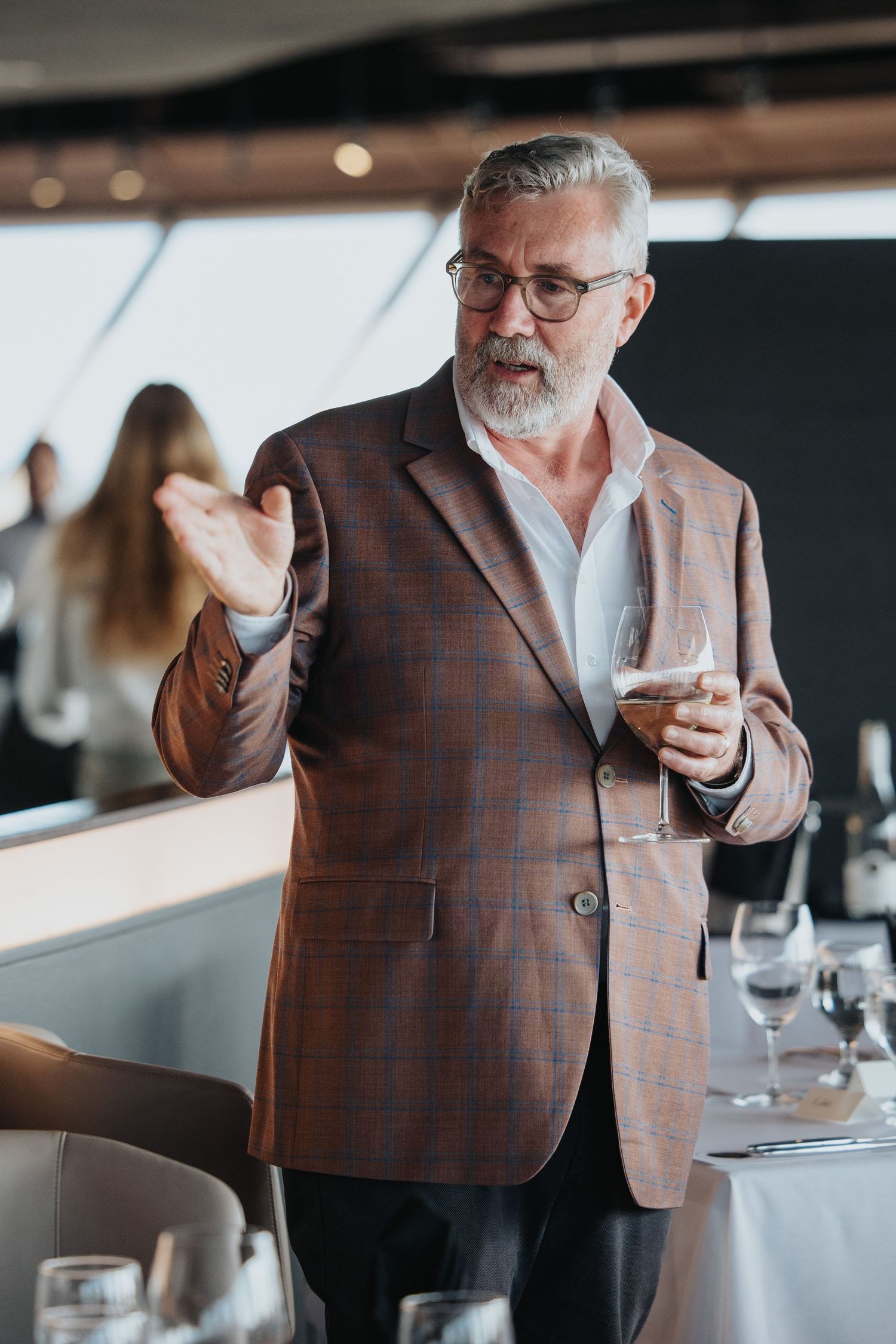 Person in brown plaid blazer gestures while holding wine glass in upscale restaurant setting.