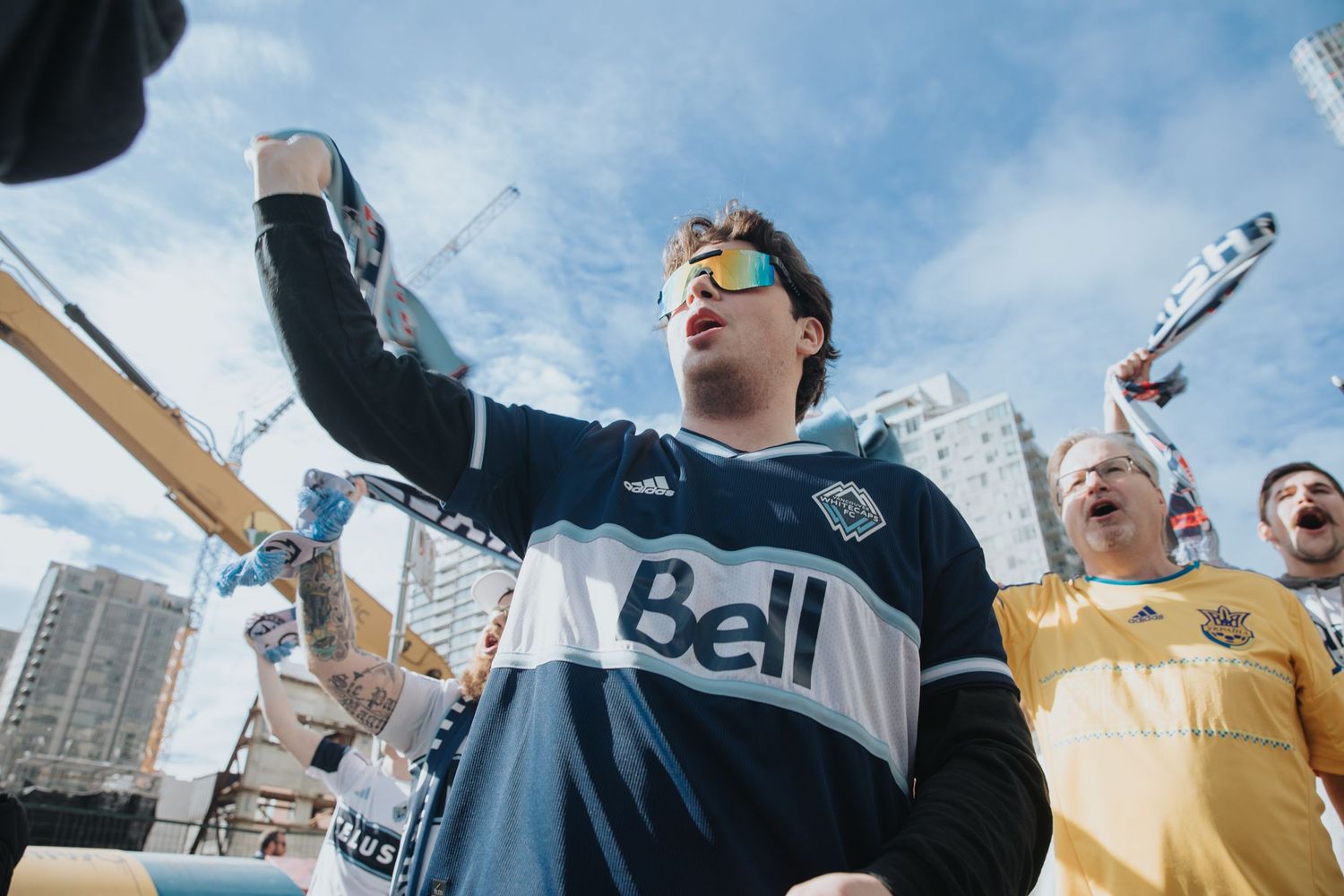 Soccer supporter in Vancouver Whitecaps jersey celebrating with arms raised against blue sky.