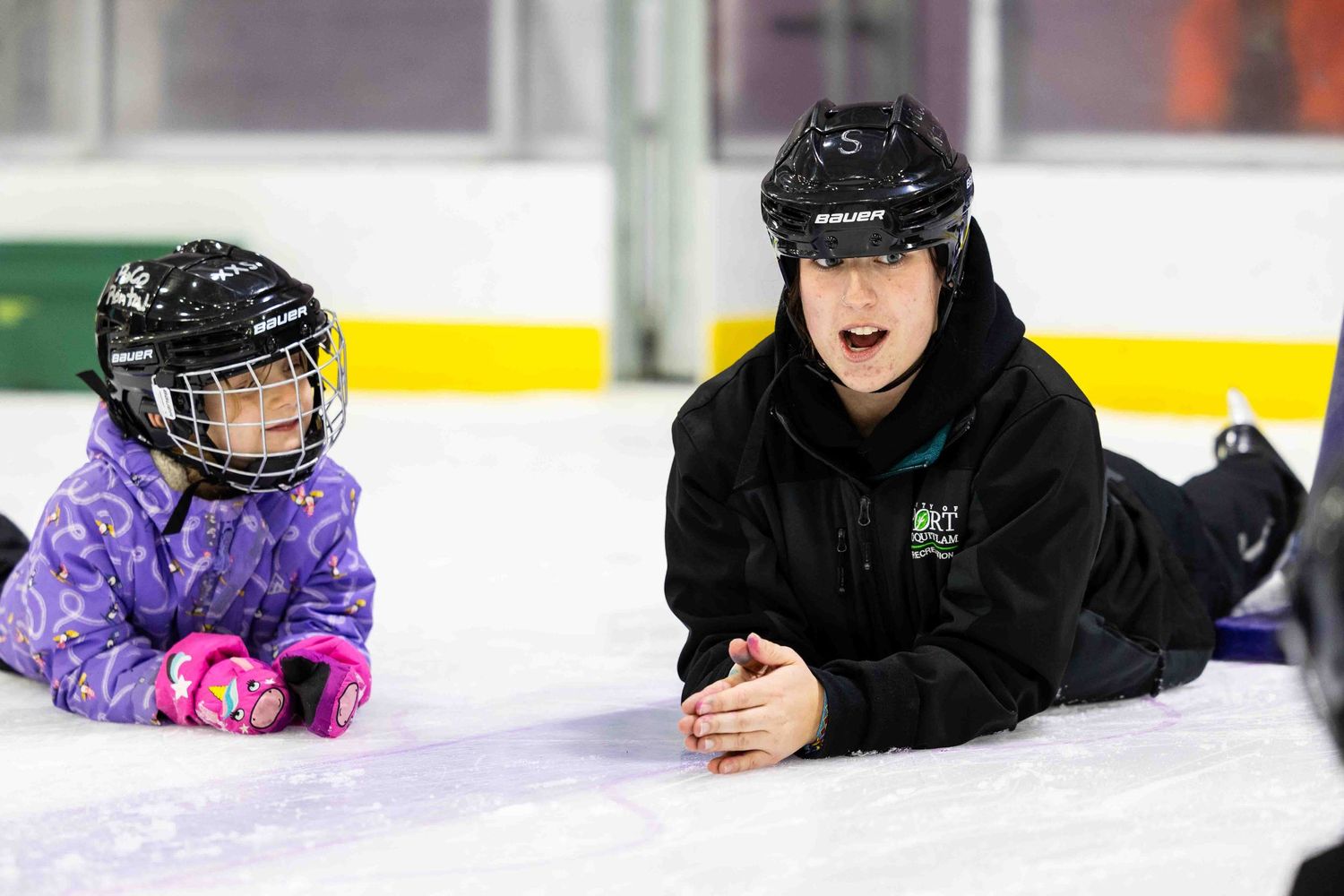 Two people in hockey helmets lying on ice rink, one in purple and one in black.