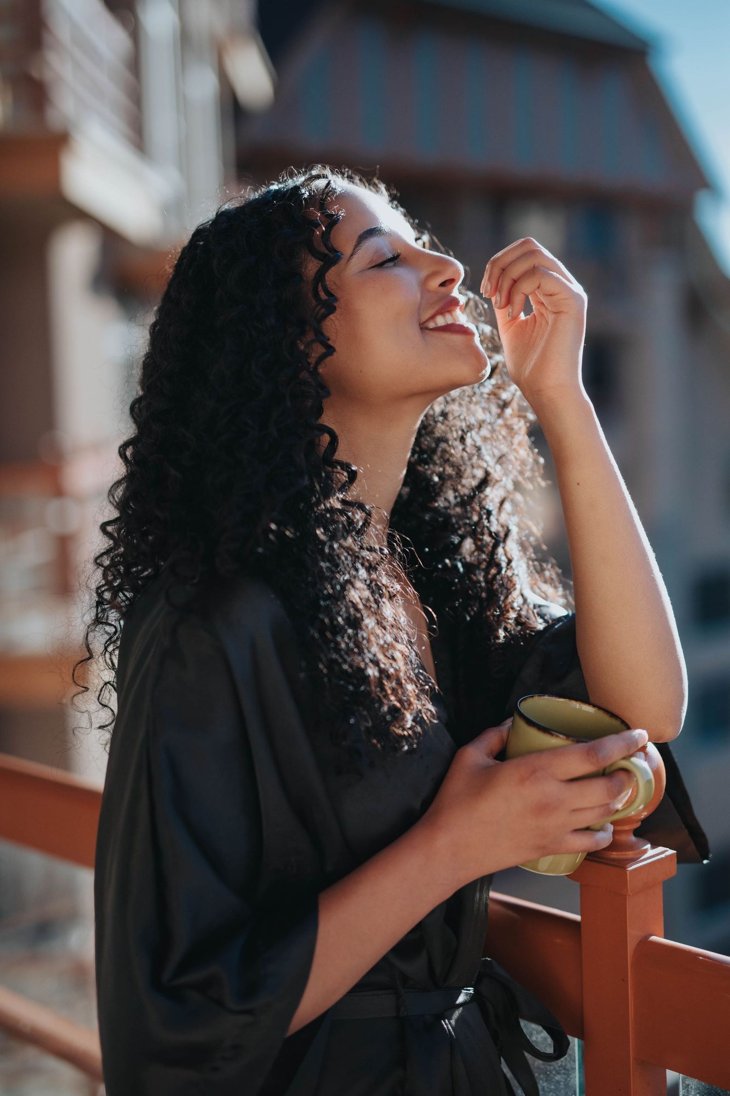 Person in black clothing enjoying a drink on a sunny balcony.