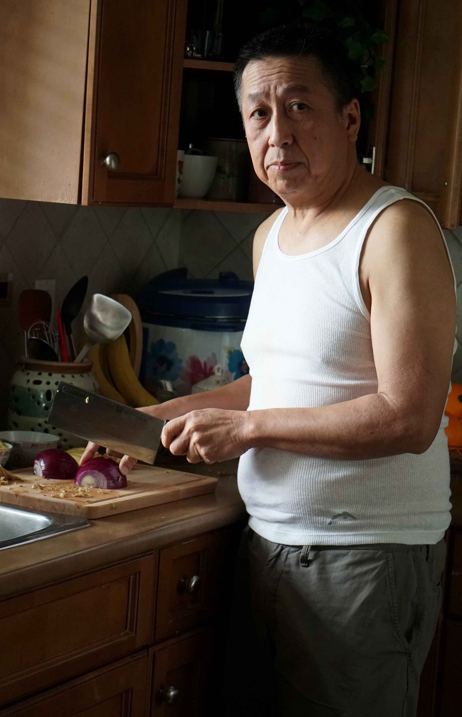 Middle-aged man in white tank top chopping red onions with cleaver in home kitchen.