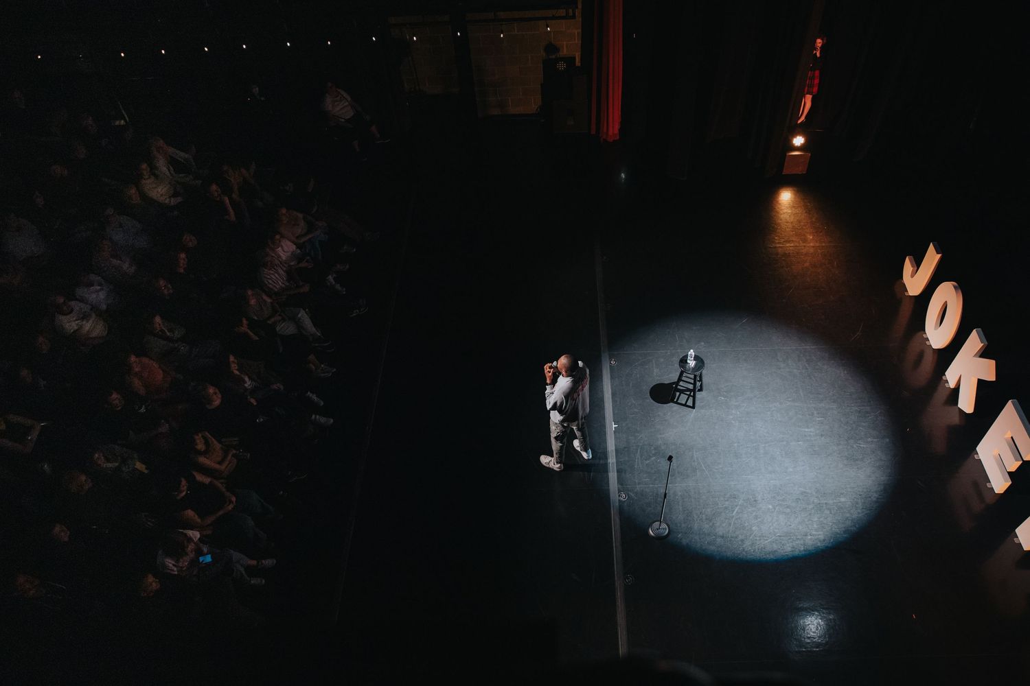 Aerial view of a comedian performing solo on a spotlight-lit stage at the Joke comedy club before a seated audience.