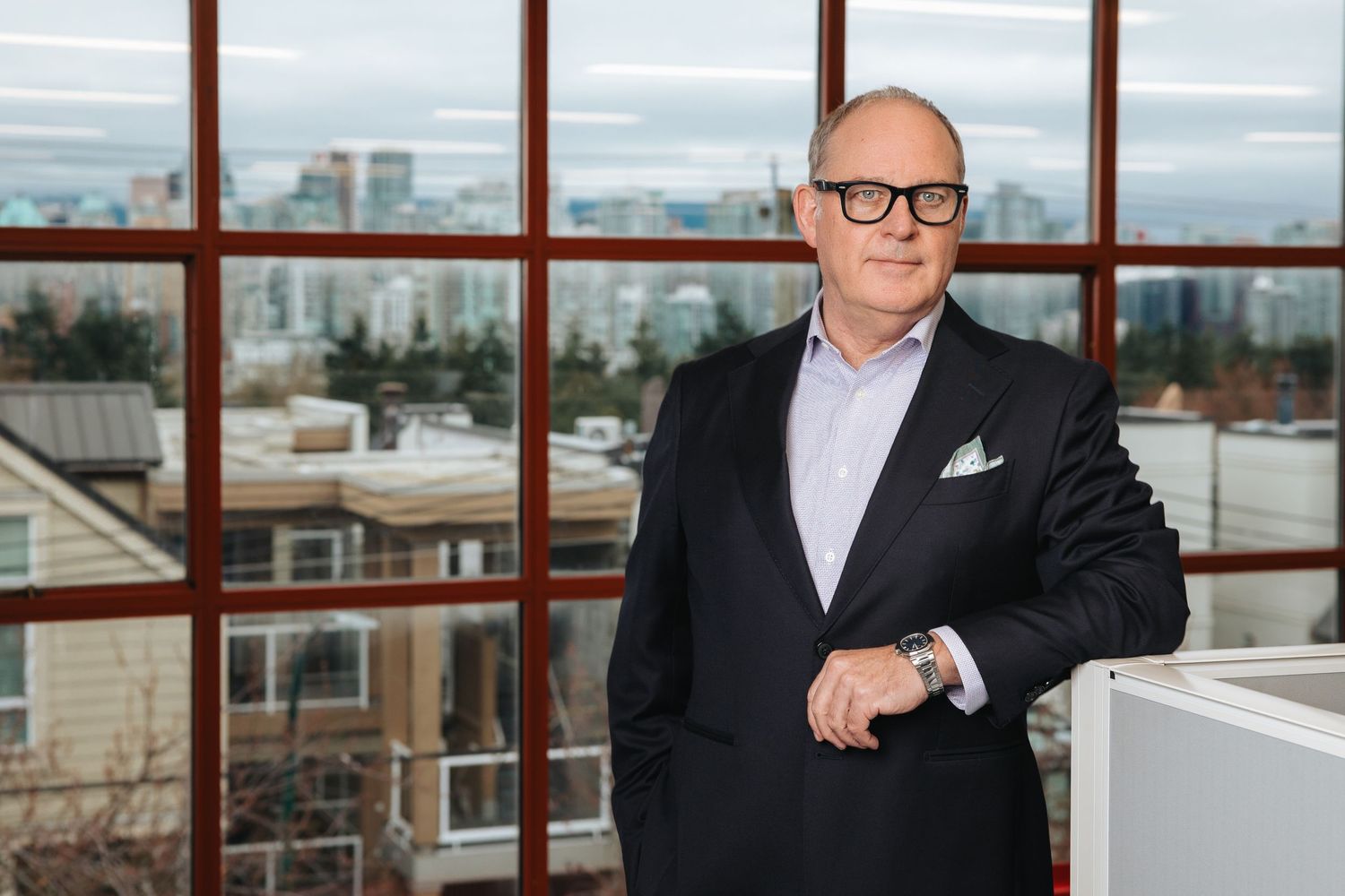 Professional in a black suit and glasses stands by large windows in a modern office building with city views.