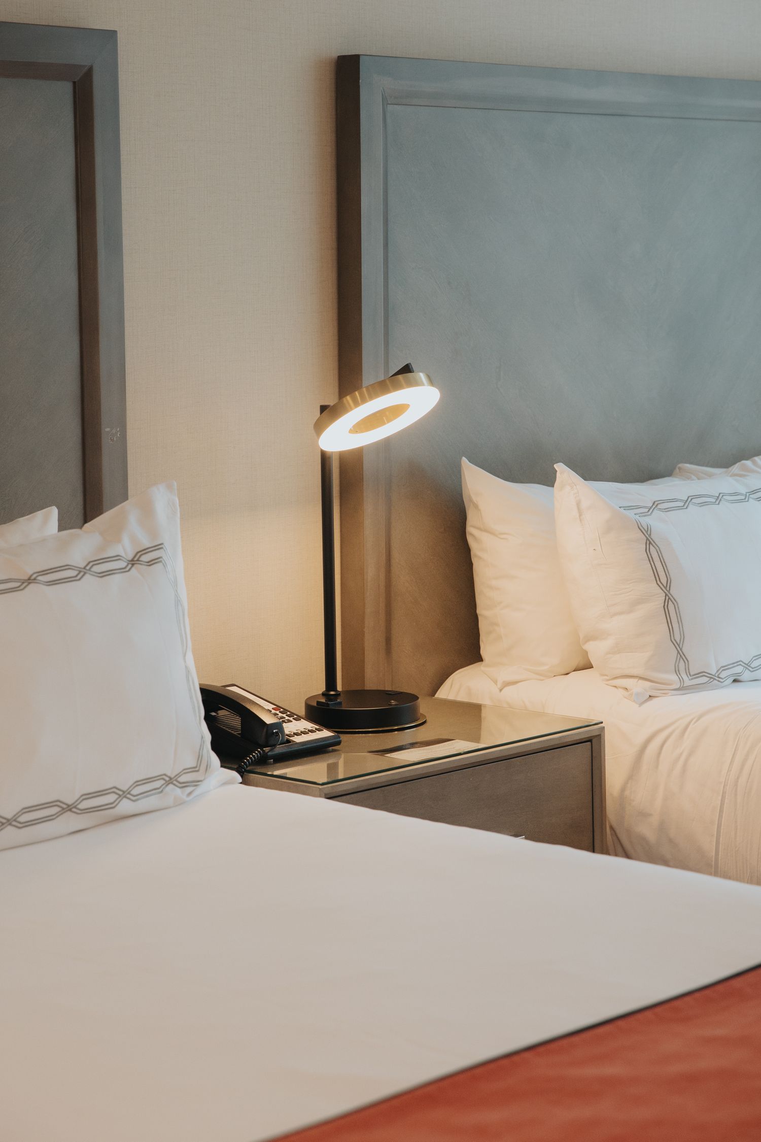Modern hotel room with two beds, white pillows, glowing bedside lamp, and telephone on nightstand.