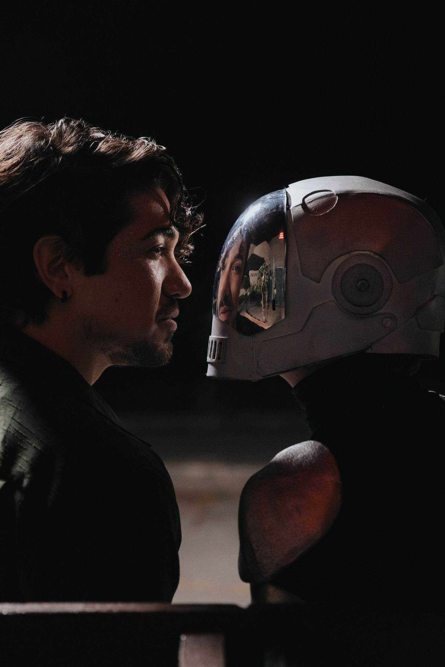 A man in profile faces a futuristic white helmet in a dramatic dark cinematic scene.