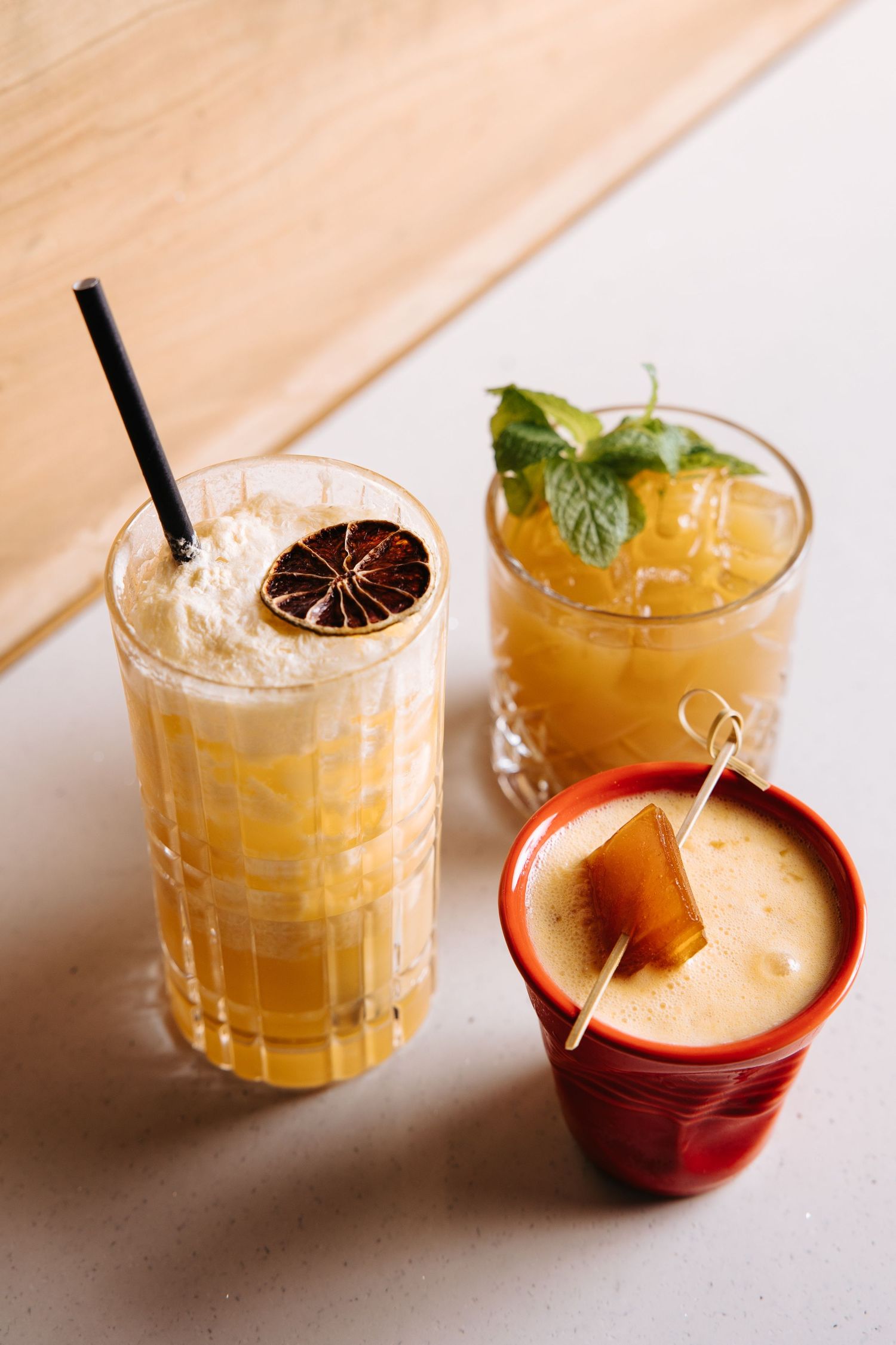 Three craft cocktails on a white surface, featuring garnishes of dried citrus, fresh mint, and a caramel cube skewer.