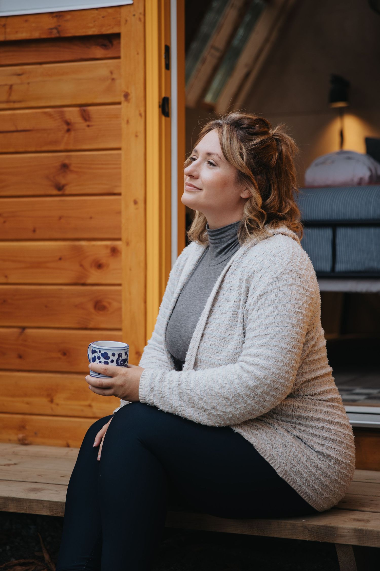 Person in grey sweater and dark pants sitting on wooden porch in relaxed pose.