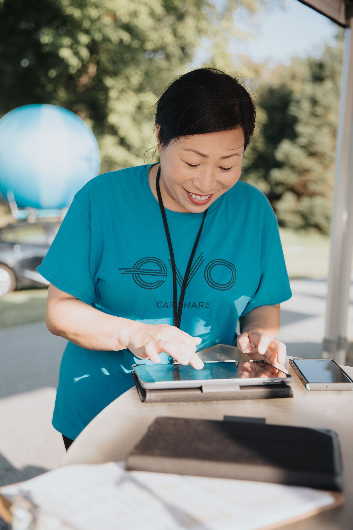 A smiling woman in a teal Evo Car Share t-shirt uses a tablet at an outdoor event booth.