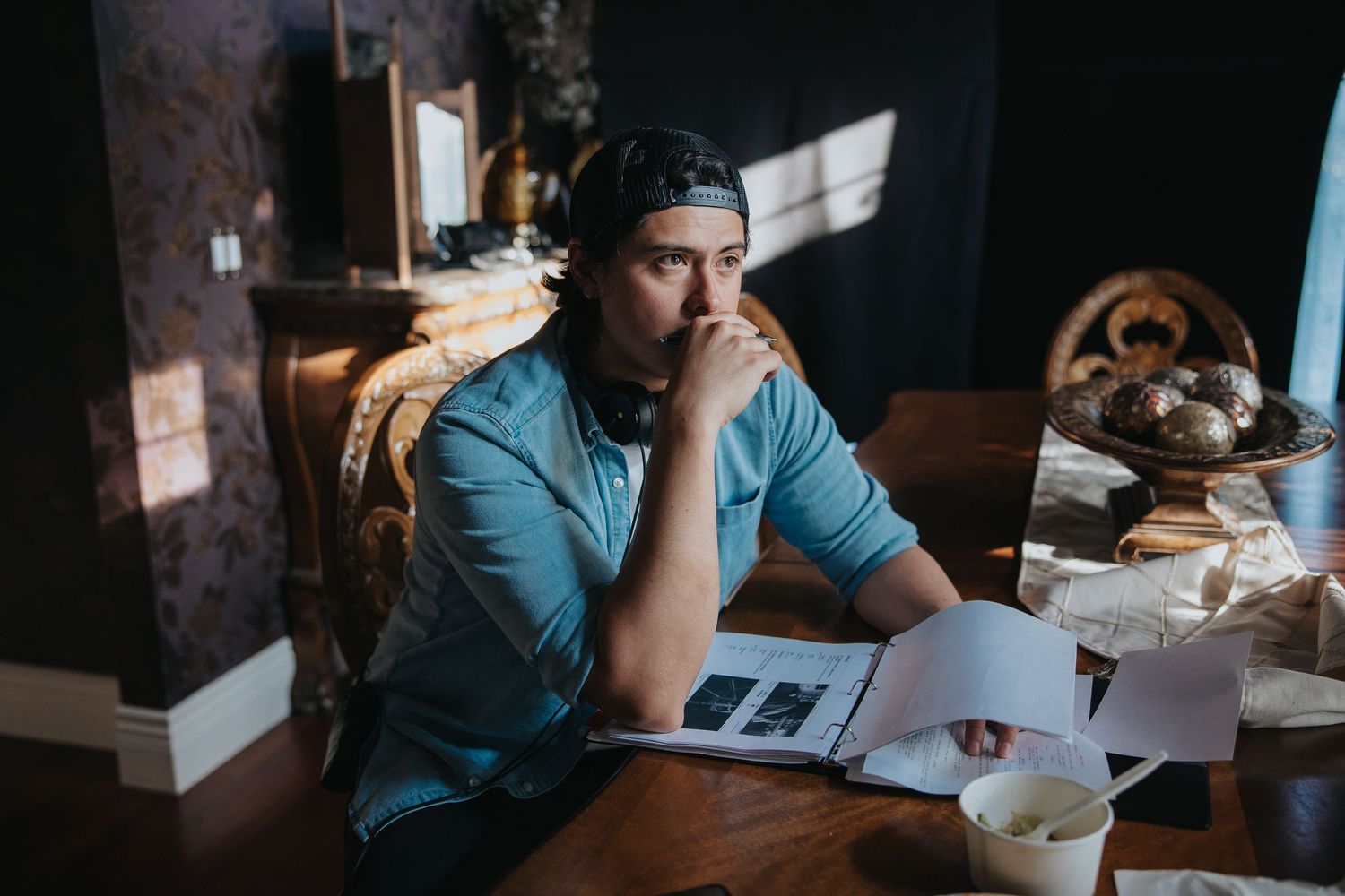 Young man in teal shirt and cap sits at wooden table reviewing documents with thoughtful expression.