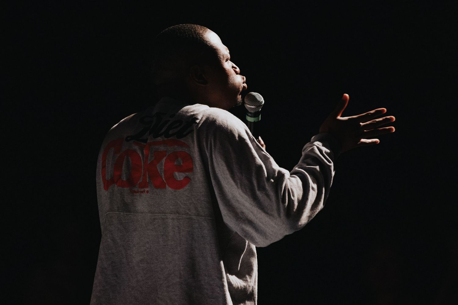 A performer in a gray Coke hoodie speaks on a dark stage, gesturing with one hand under dramatic lighting.