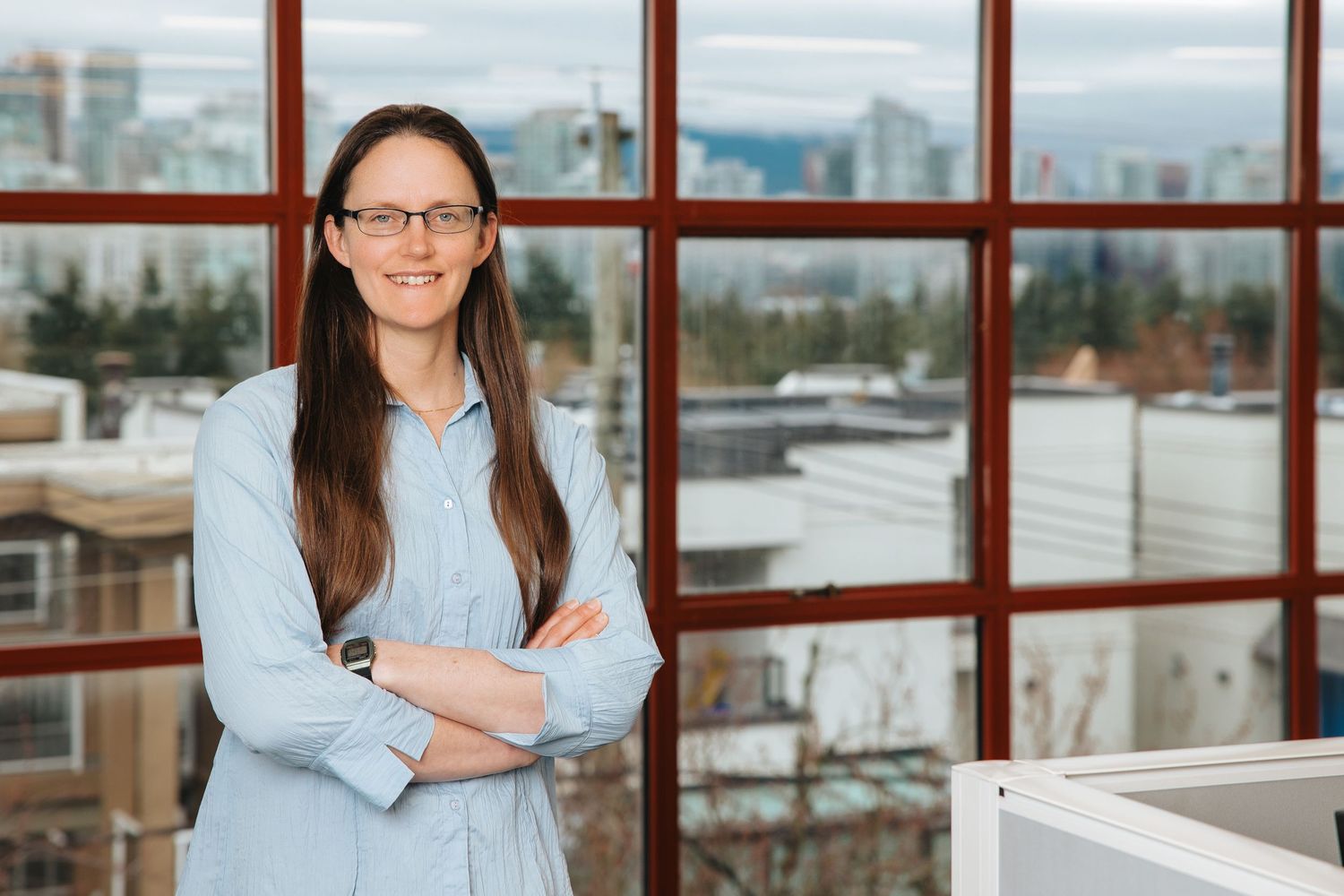 Professional in glasses and light blue shirt stands with arms crossed by office windows with city view.