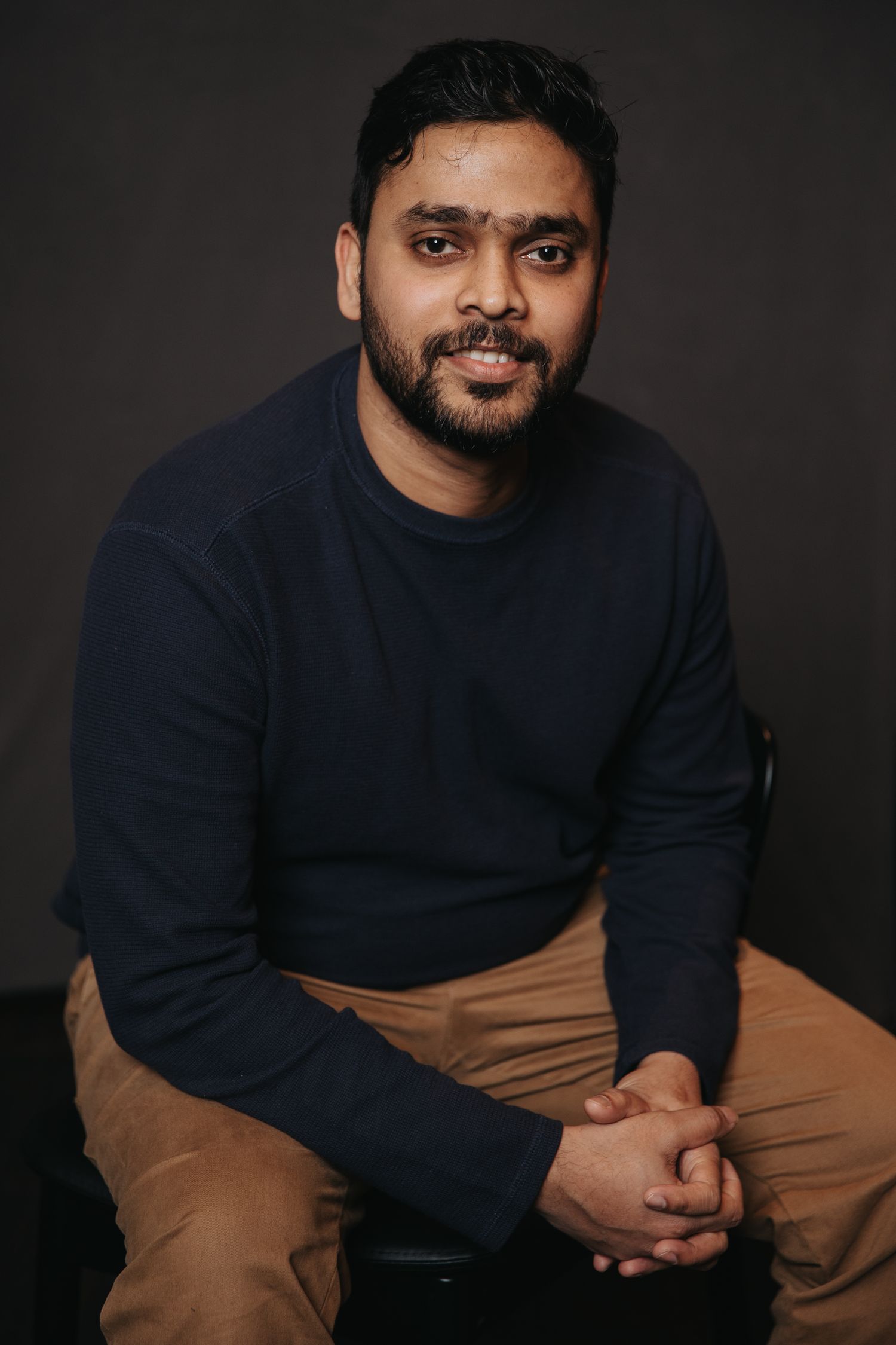 Portrait of person in navy sweater and khaki pants seated against dark backdrop.
