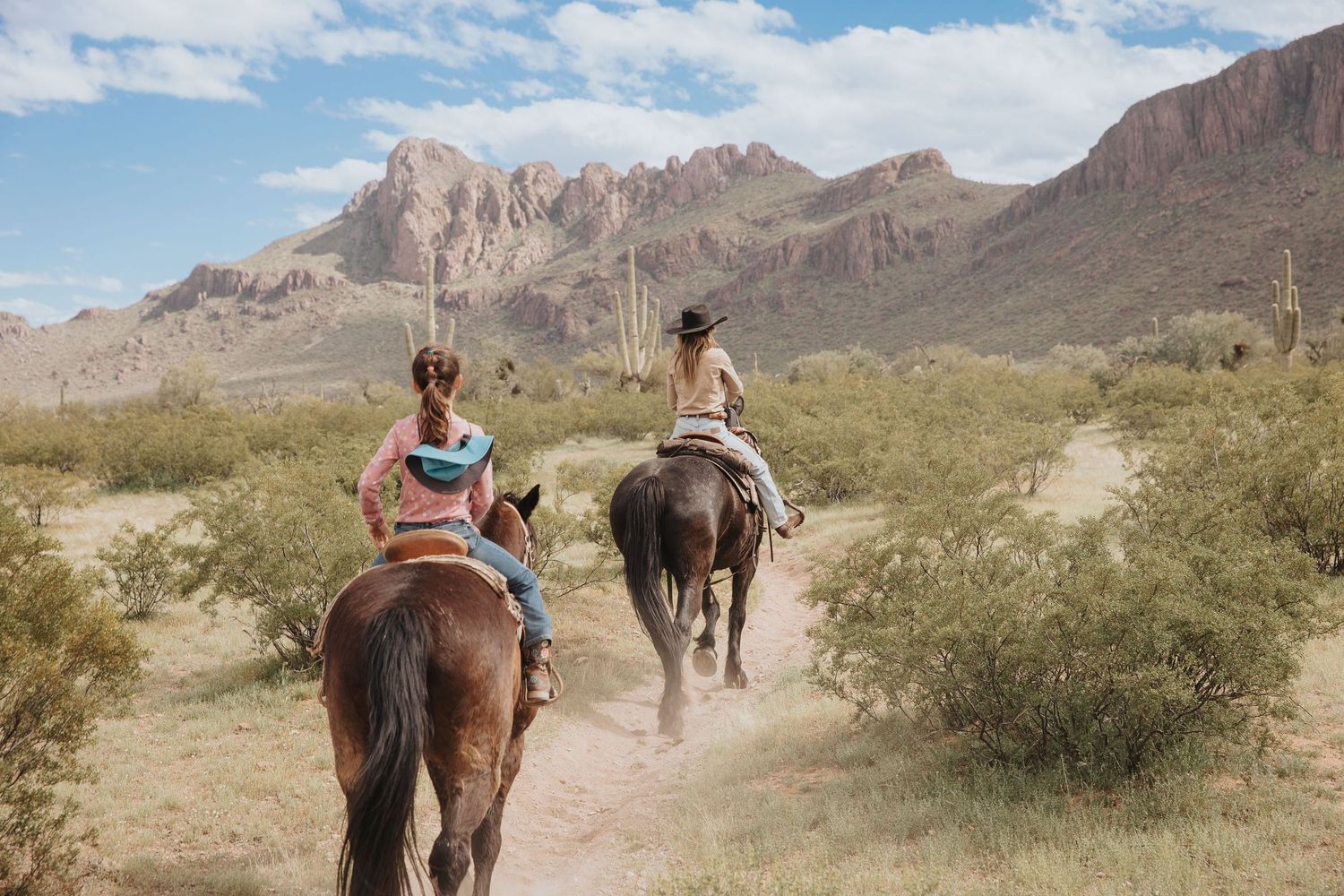Horseback riders traversing a desert trail with rugged mountain peaks in the background.