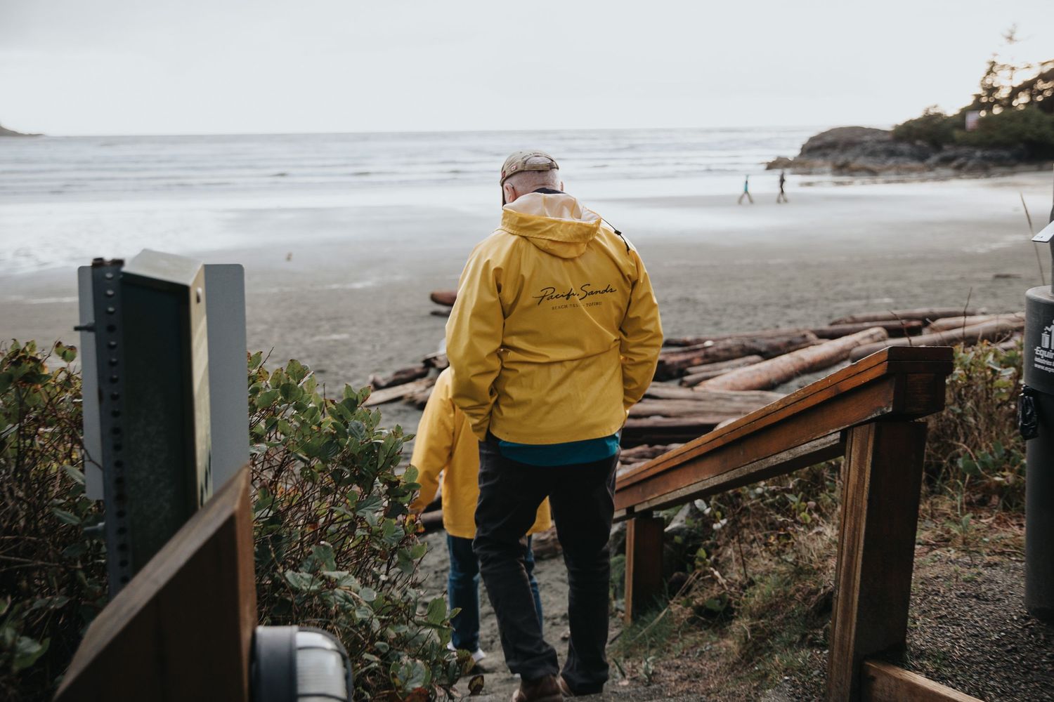 Two people in yellow raincoats walking along a rugged coastal path with driftwood.