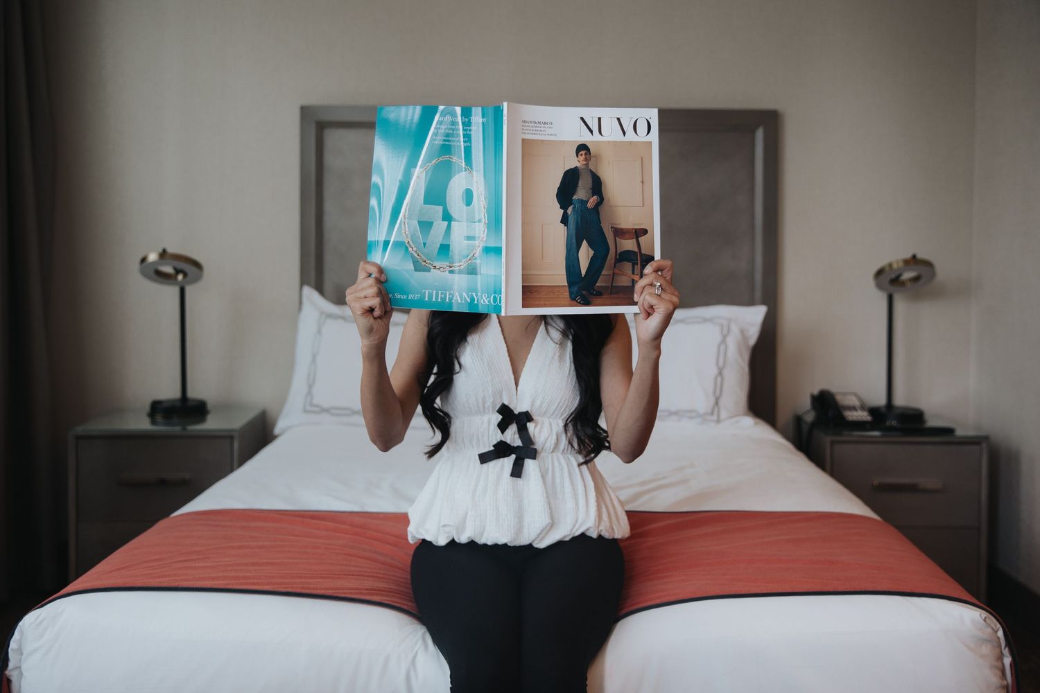 Woman reading a magazine on a neatly made hotel bed with white linens and twin nightstands.