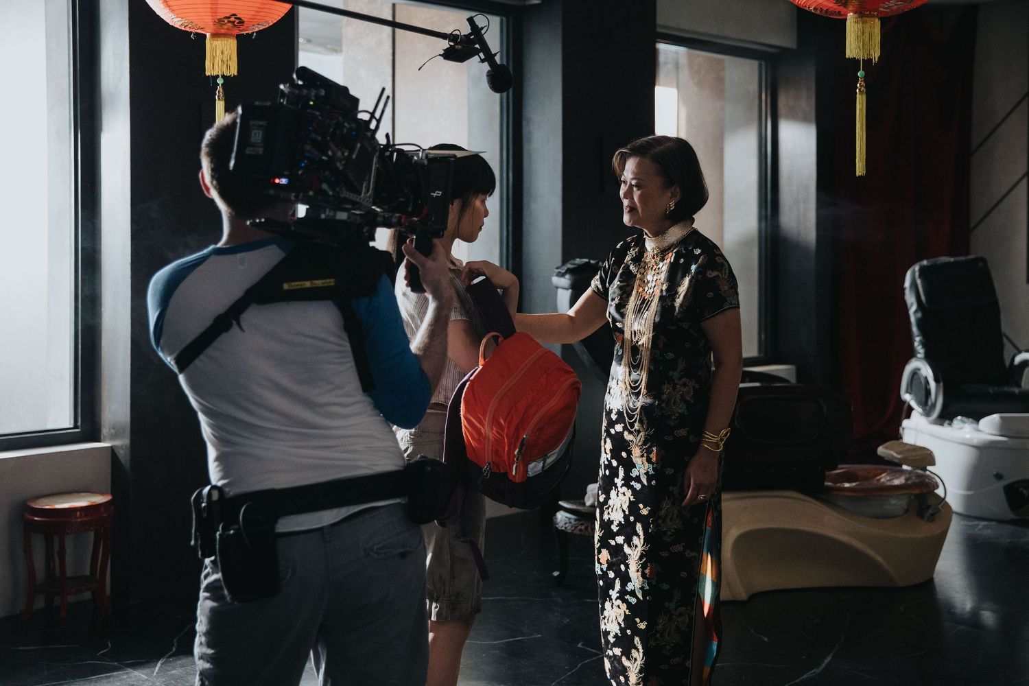 Film crew interviewing an elderly woman in a traditional Chinese qipao dress in a decorated Asian restaurant.