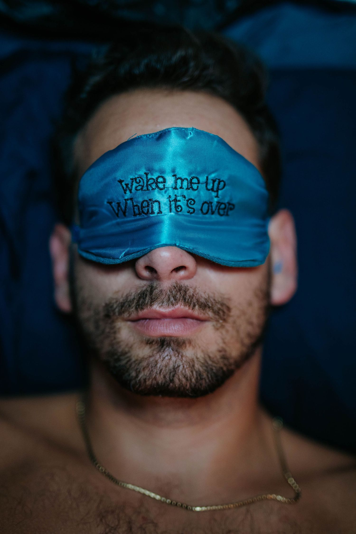 Man wearing blue satin sleep mask embroidered with wake me up when its over lying in bed.
