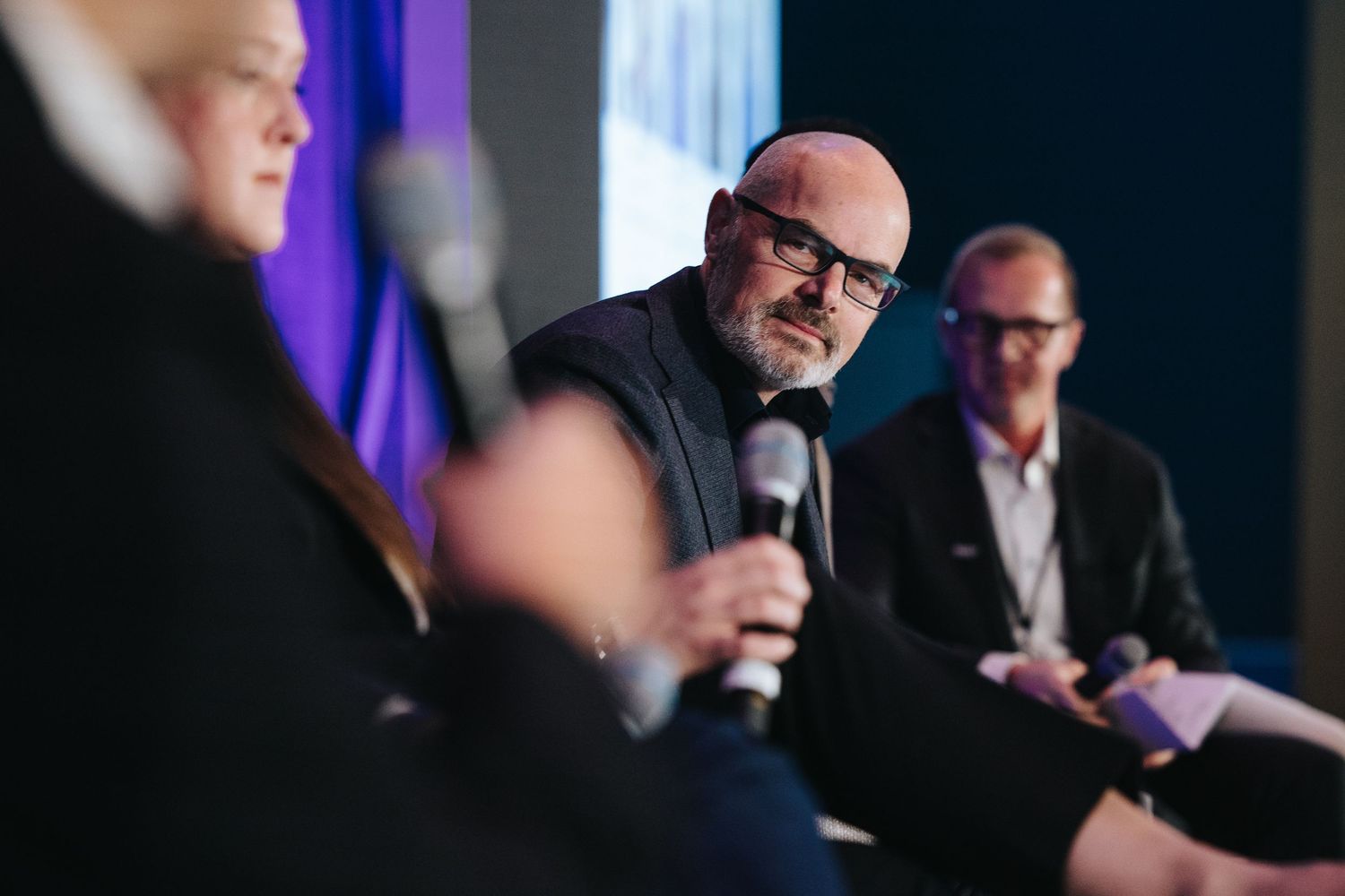 Business executives participate in a panel discussion on stage with microphones and purple lighting.