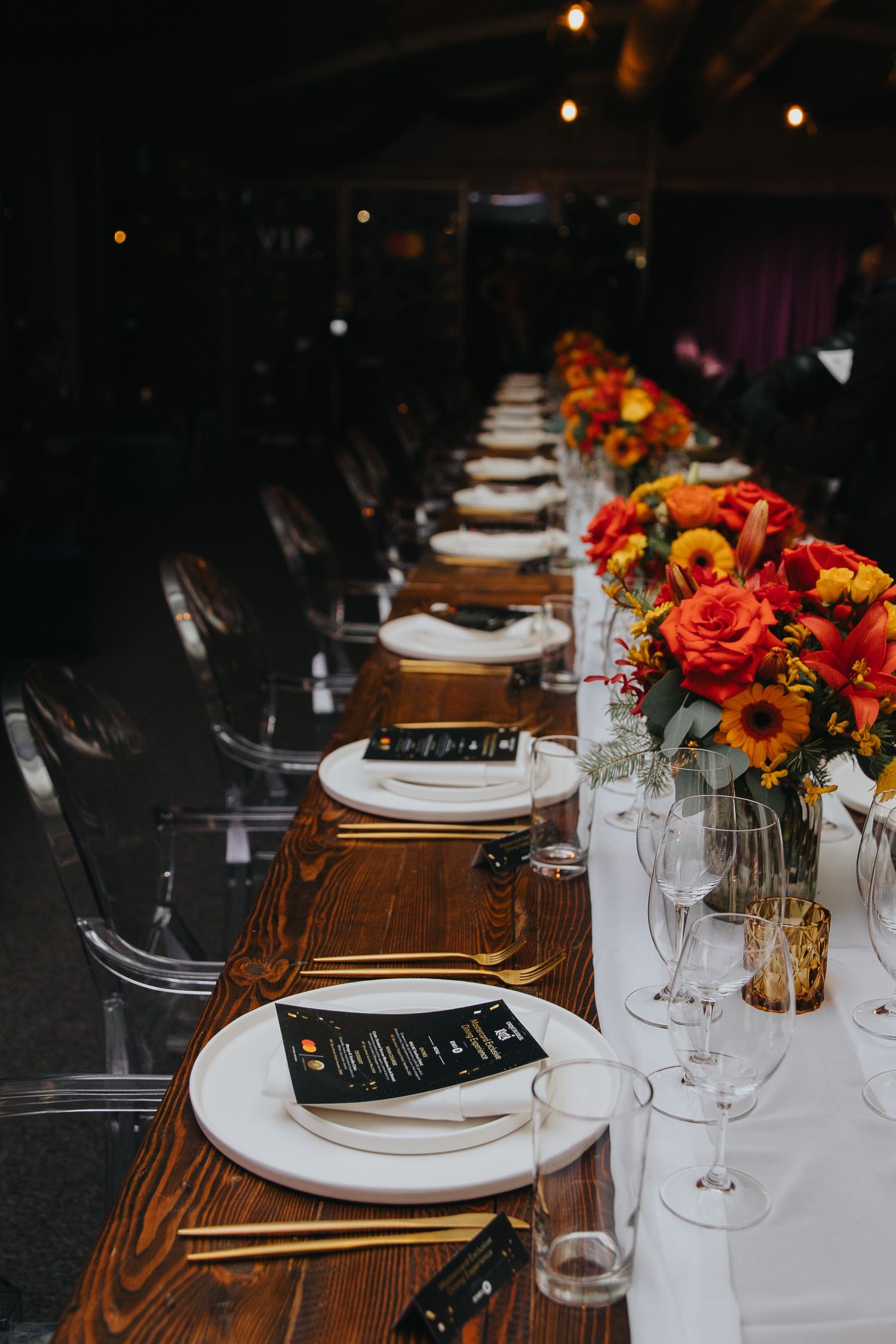 Elegant table setting with white plates and vibrant orange floral centerpieces on a dark wooden table.
