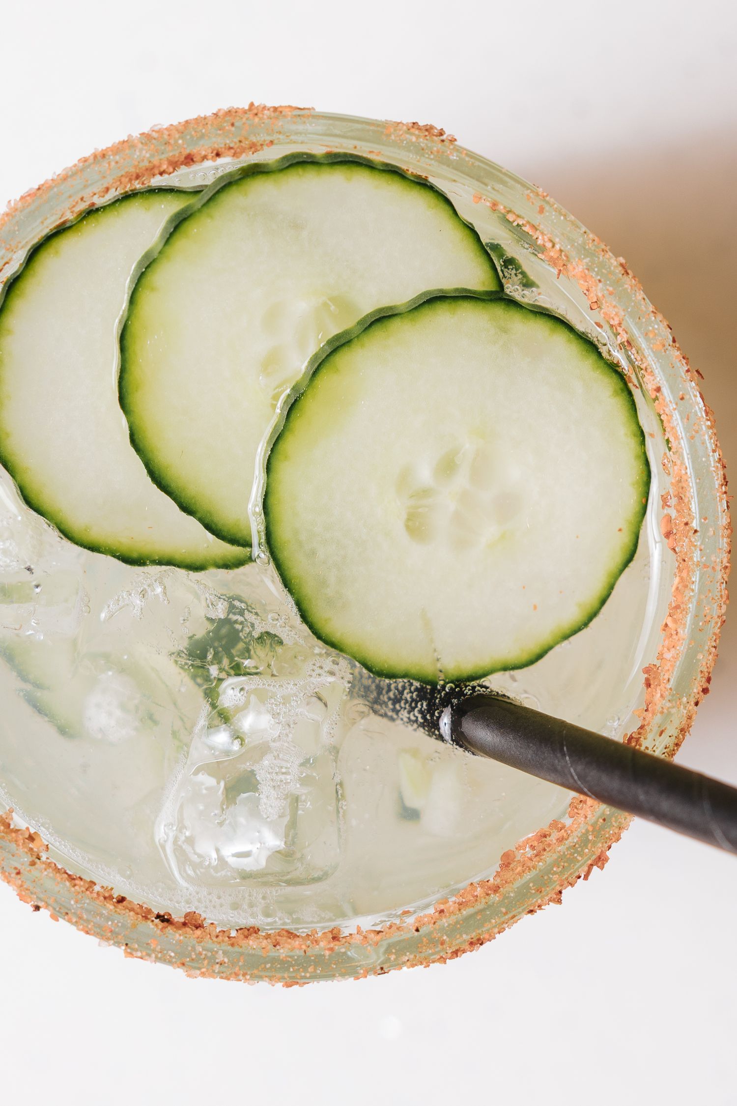 Overhead view of a cucumber margarita with ice, black straw, and spiced salt rim garnished with fresh cucumber slices.