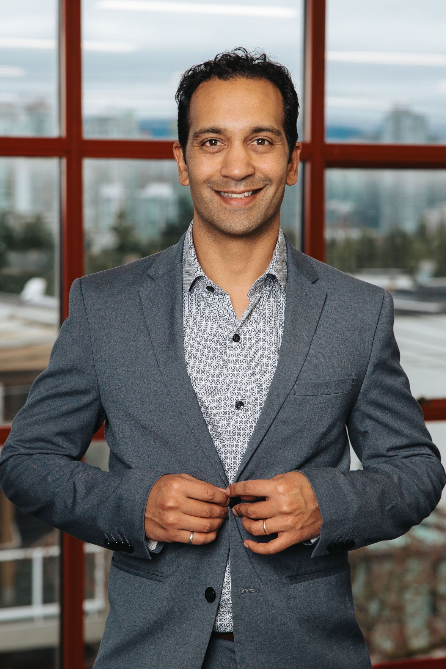 Professional wearing a grey suit jacket and patterned shirt adjusts his attire in a modern office setting with windows.