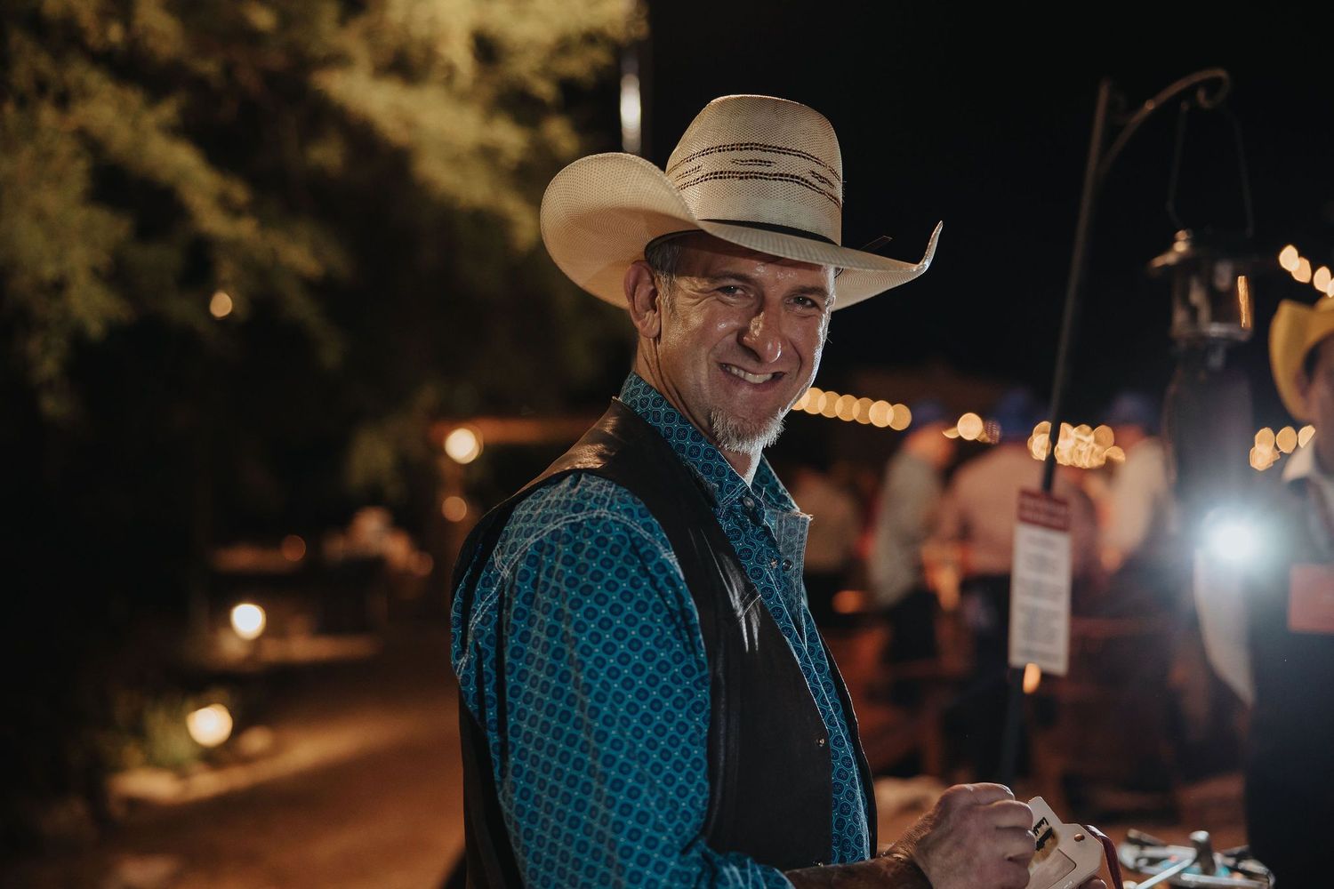 A smiling man wearing a white cowboy hat and plaid shirt at an outdoor evening event.