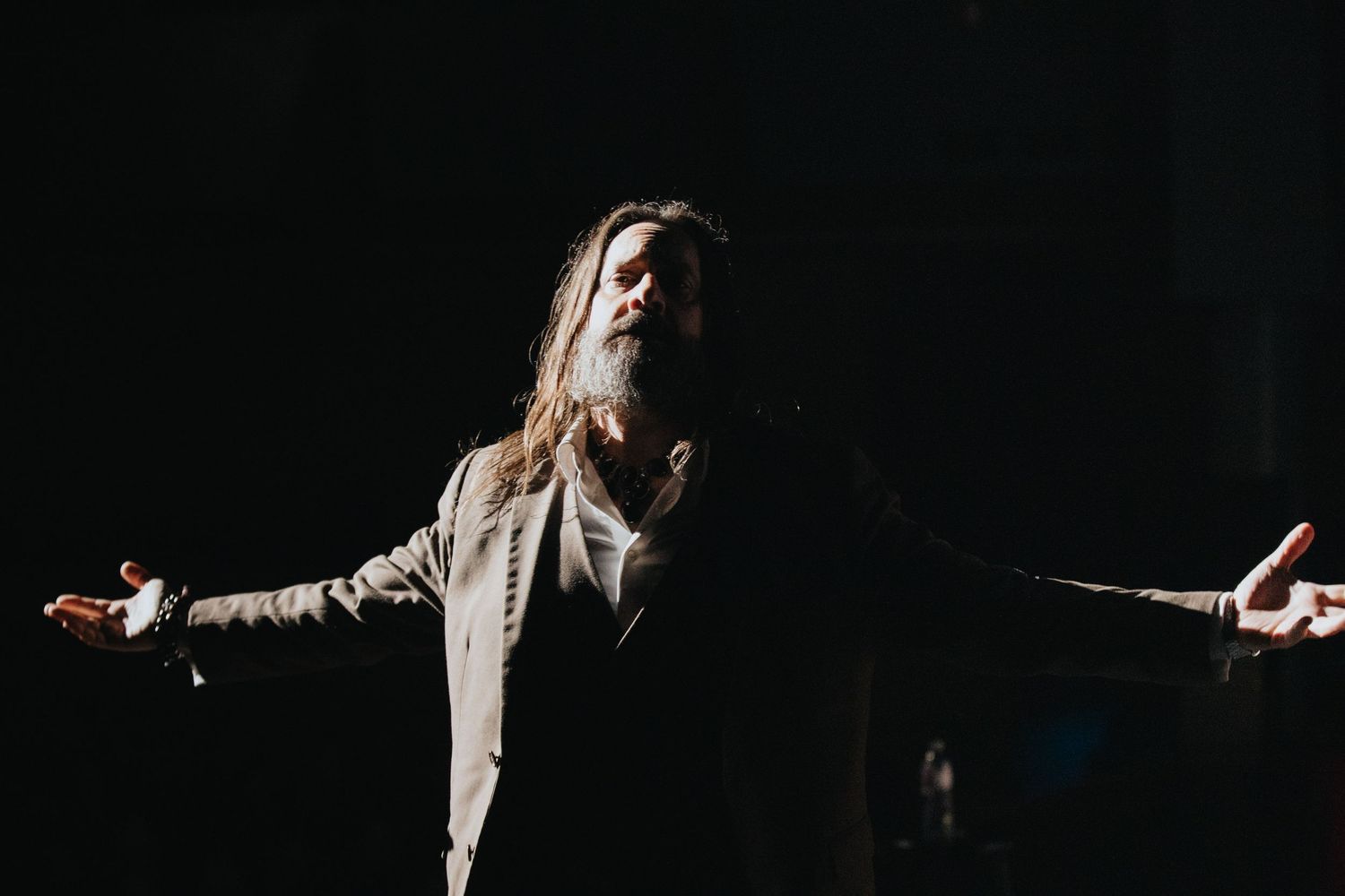 Long-haired bearded man in light jacket performing on dark stage with arms outstretched dramatically.