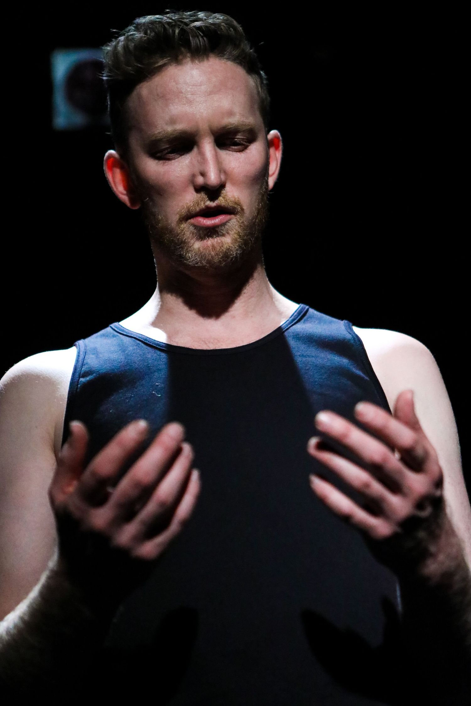 Male performer in dark tank top gesturing with both hands on dimly lit stage.