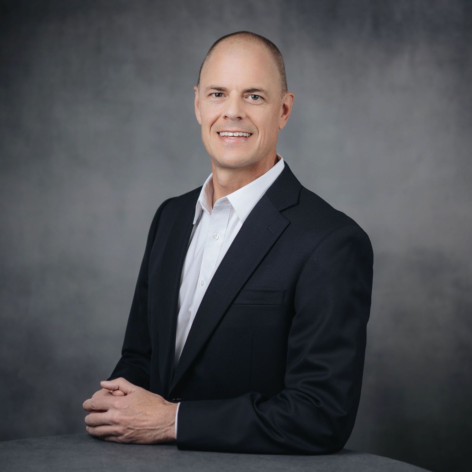 Professional headshot of someone in a black suit against a grey backdrop.