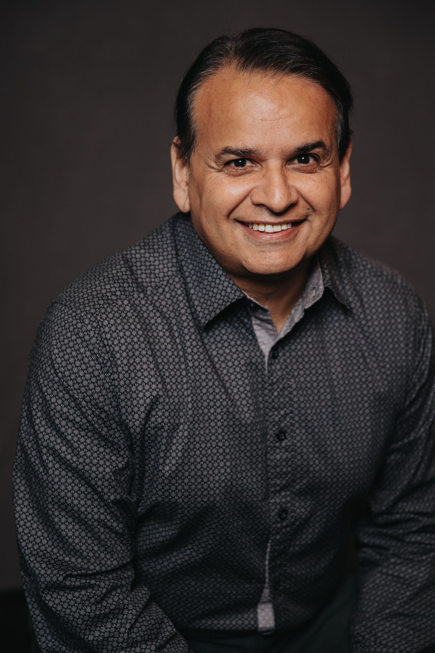 Professional headshot of person wearing gray patterned button-down shirt against dark background.